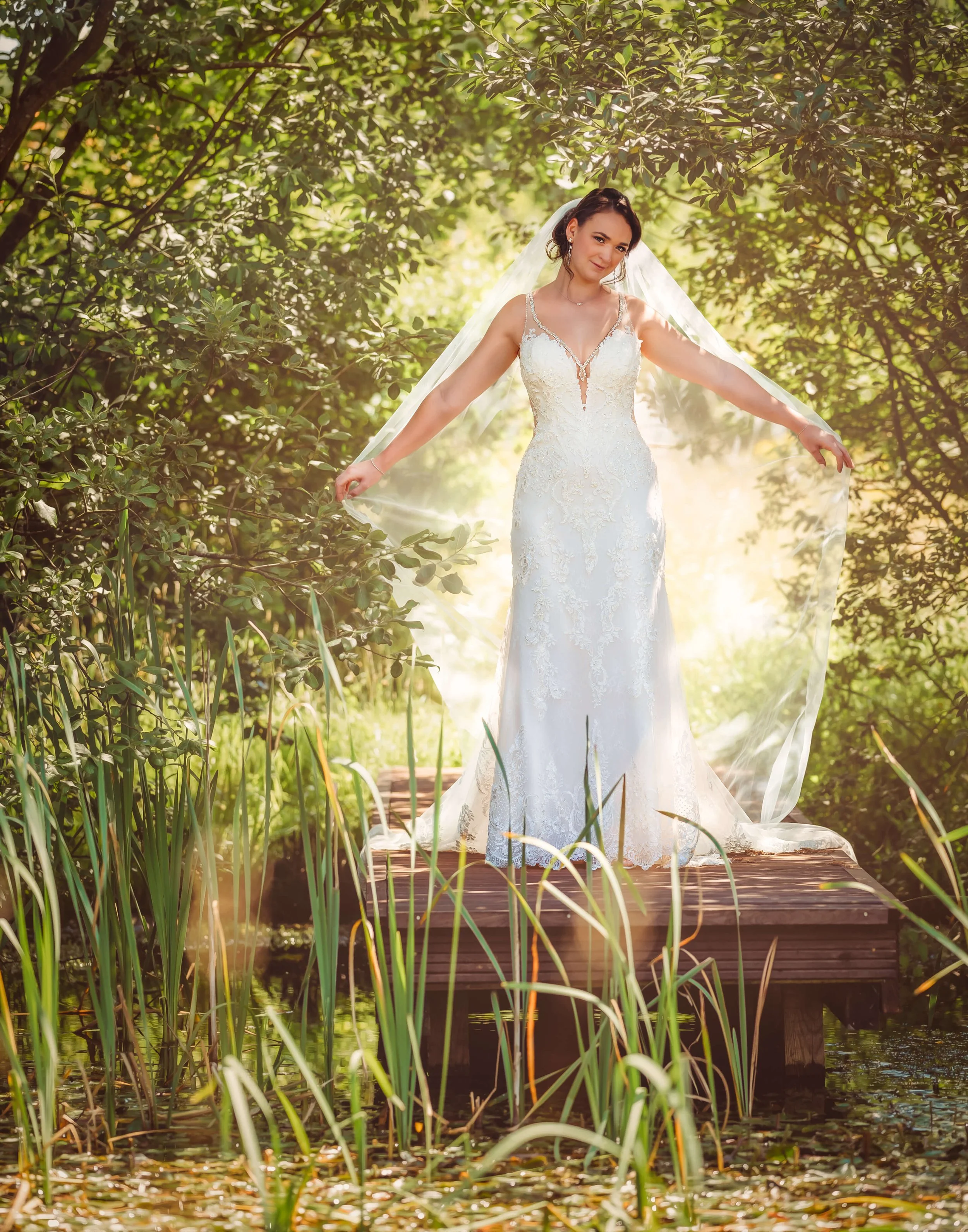 A bride in a white wedding dress standing on a wooden dock surrounded by lush green trees and water with tall reeds, holding her veil out to the sides.