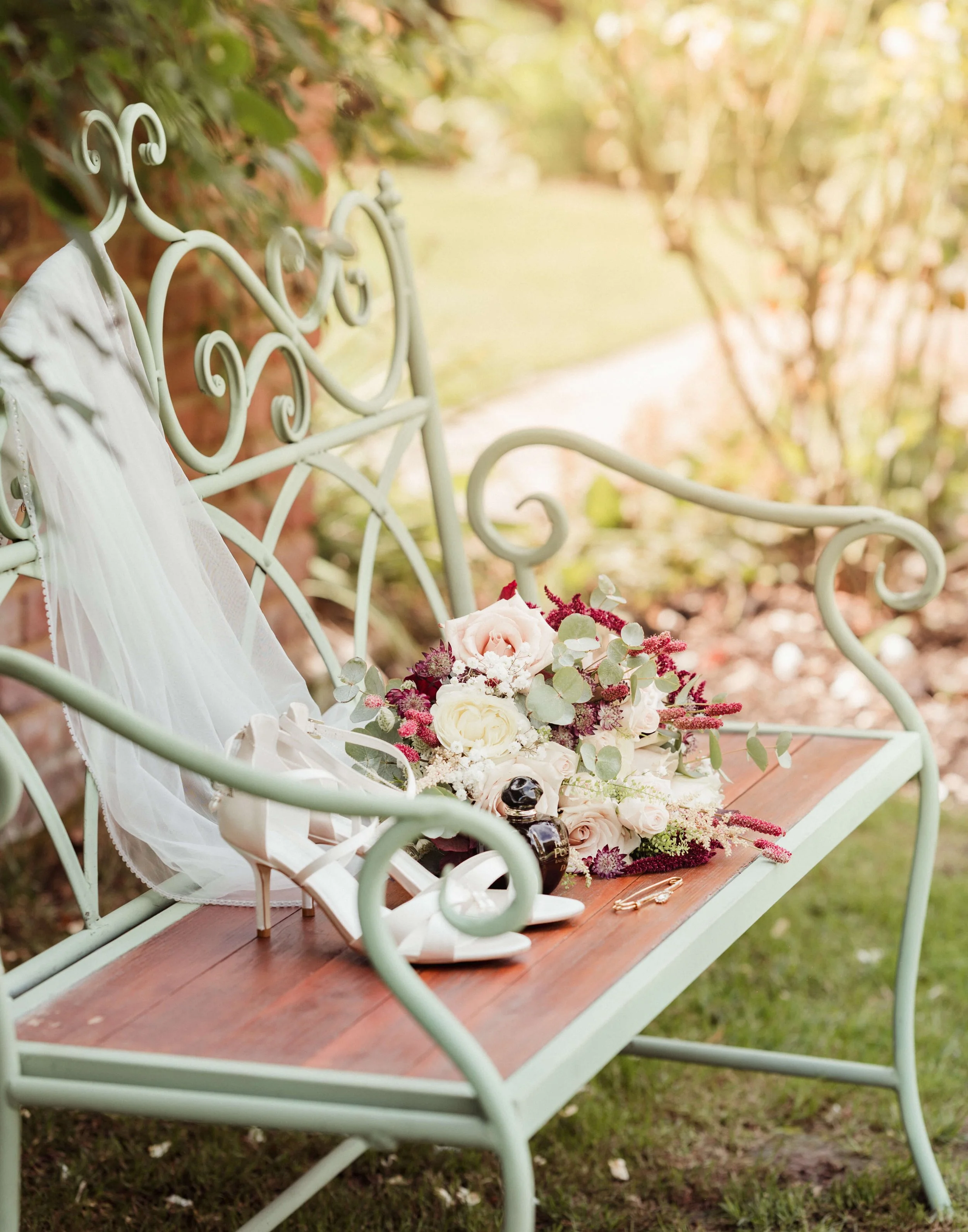 A vintage green garden bench with a wooden seat, decorated with a white veil, a bouquet of pink and white roses, and a pair of white high-heeled shoes, set outdoors on grass near a brick wall and plants.