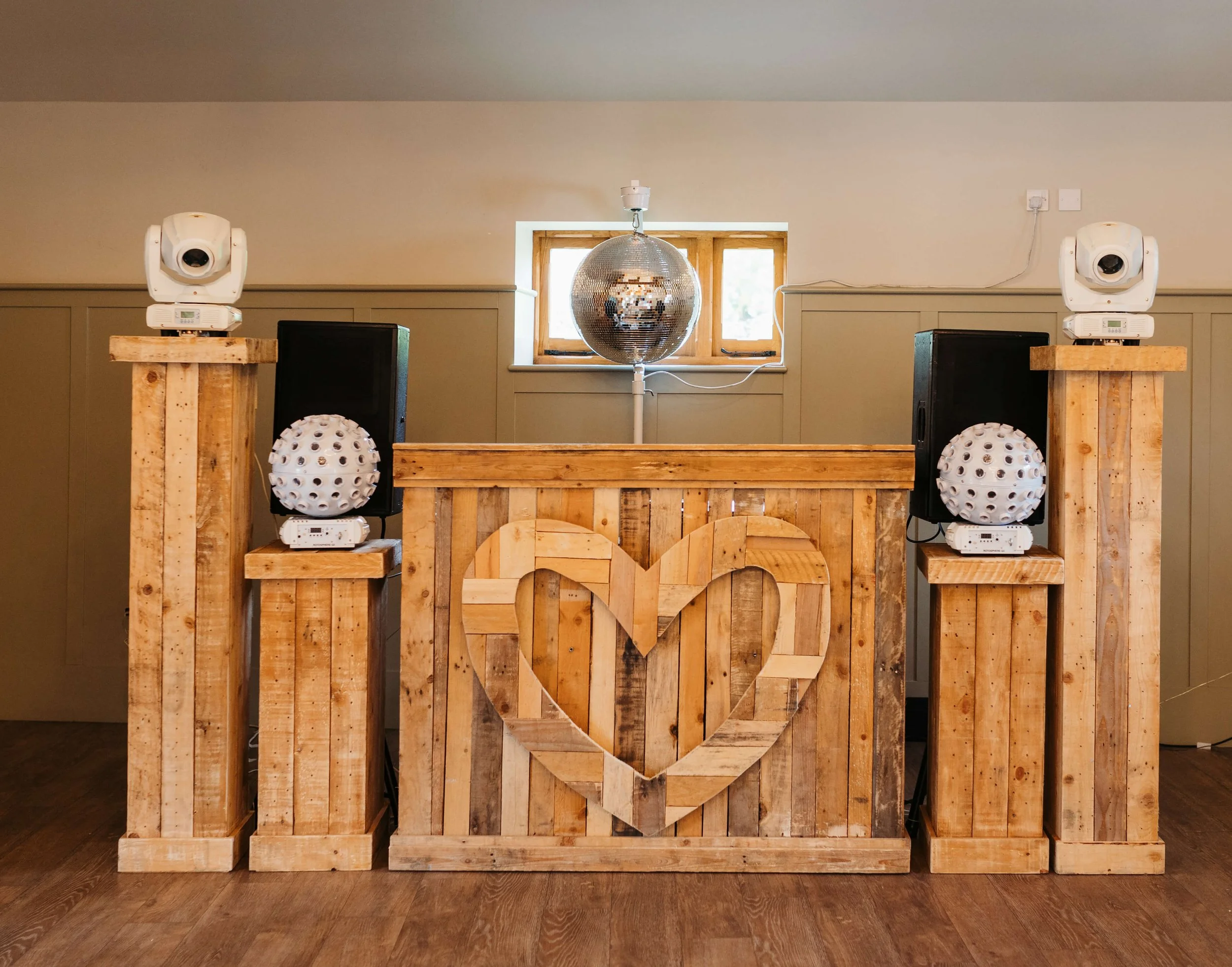 A rustic wooden DJ booth with two tall wooden pedestals on each side, each holding a white spotlight and a white spherical speaker. In front of the booth is a wooden heart decoration. Behind the booth, a window with two small panes is visible, with a