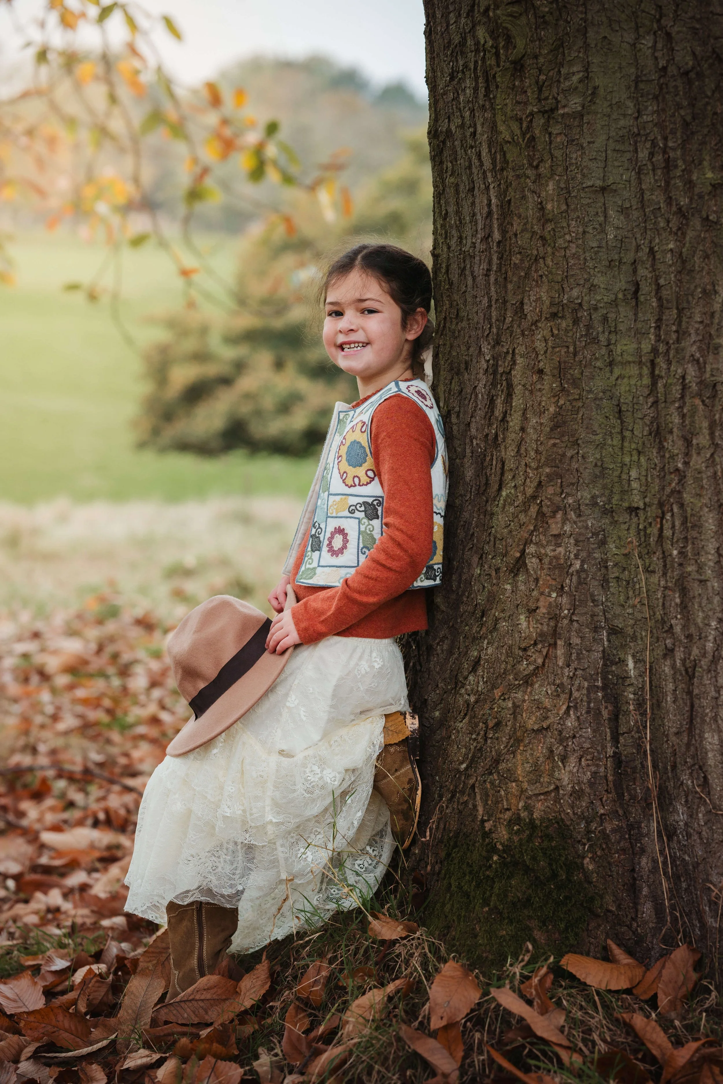 A young girl leaning against a tree outdoors during autumn, holding a tan hat, with fallen leaves on the ground, smiling at the camera.