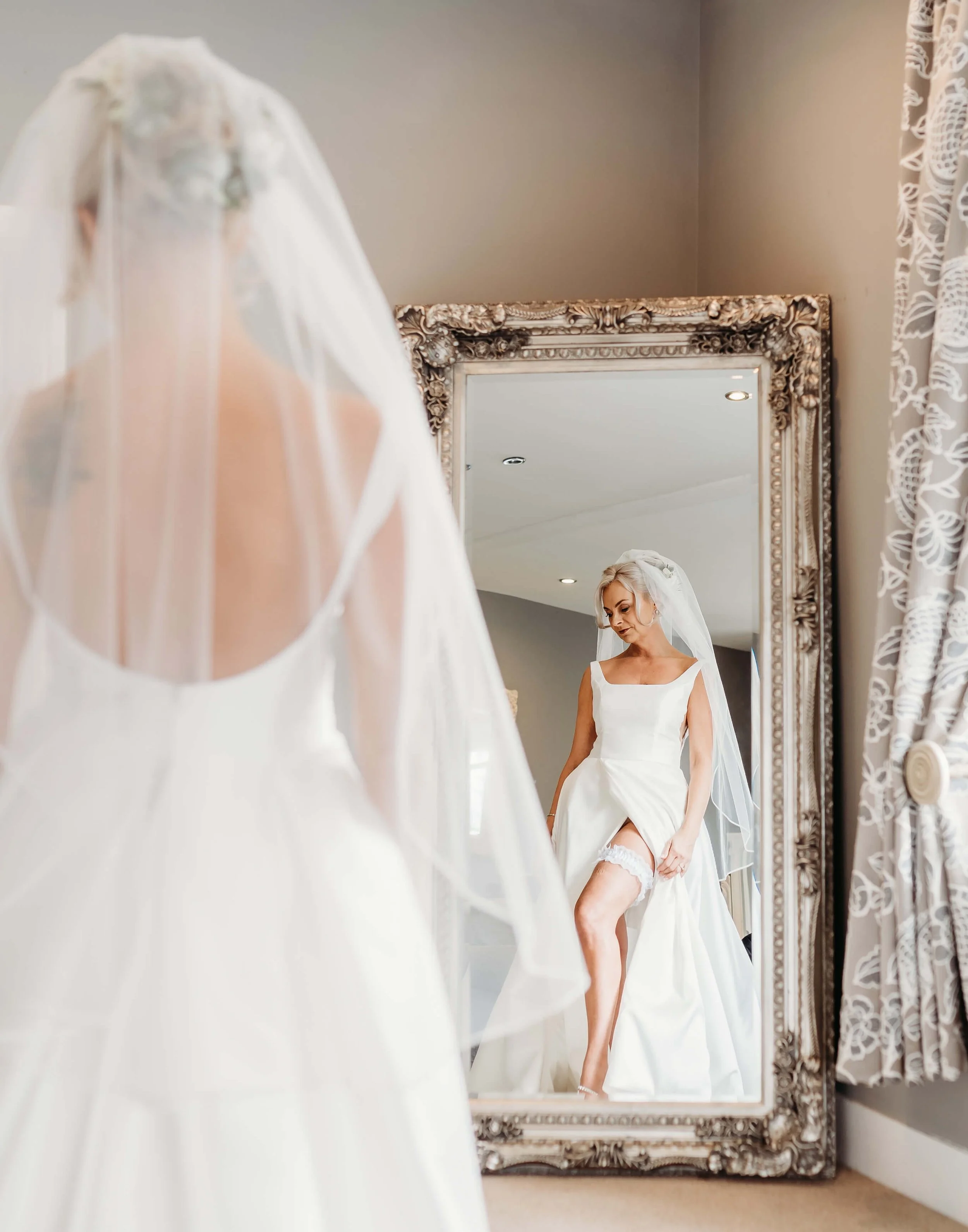 A bride in a white wedding dress looking at her reflection in an ornate mirror, adjusting her garter.