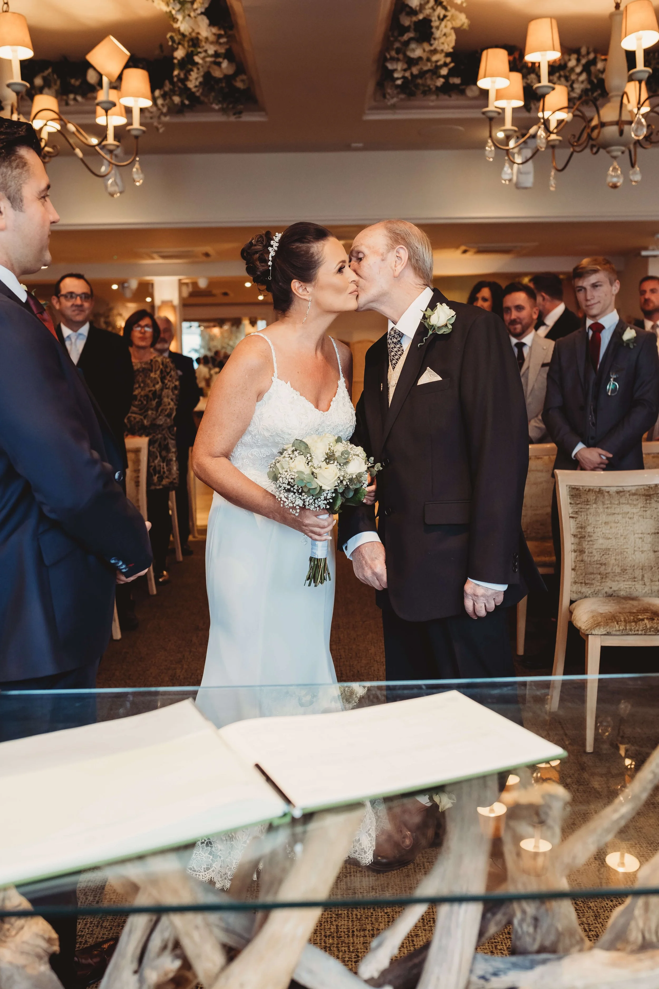 Bride and elderly man sharing a kiss at a wedding ceremony, with wedding guests in the background.