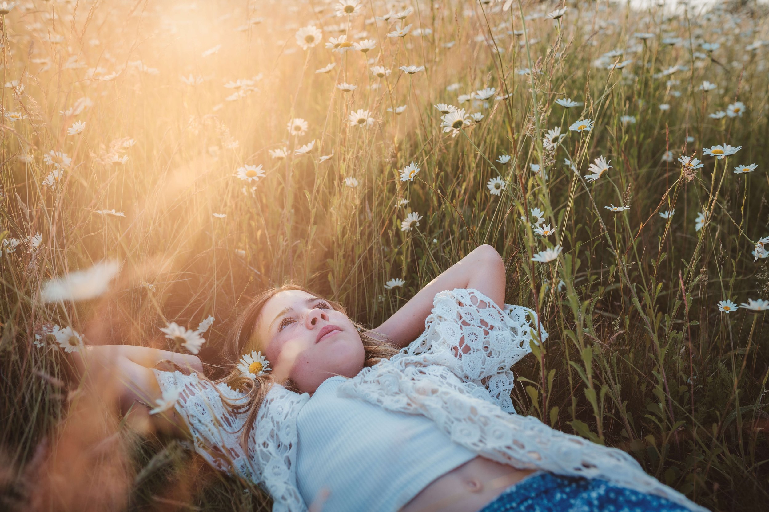 A young girl lying in a field of daisies during sunset, looking up with her hands behind her head.