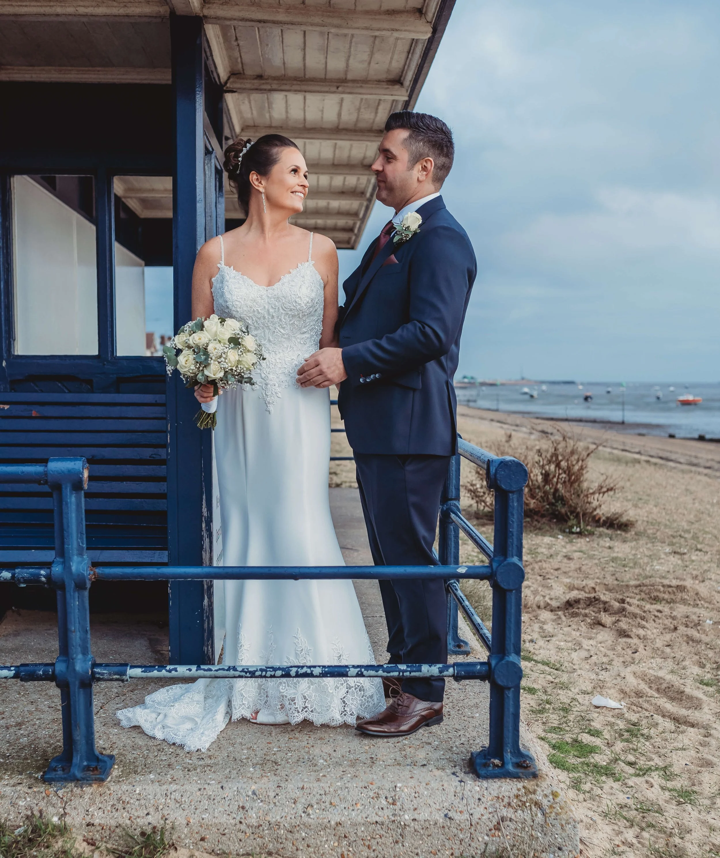 A bride and groom stand close together on a beachside terrace, gazing into each other's eyes. The bride holds a bouquet of white roses, and the groom wears a navy suit. A building with blue trim is behind them, and the ocean with boats is visible in 