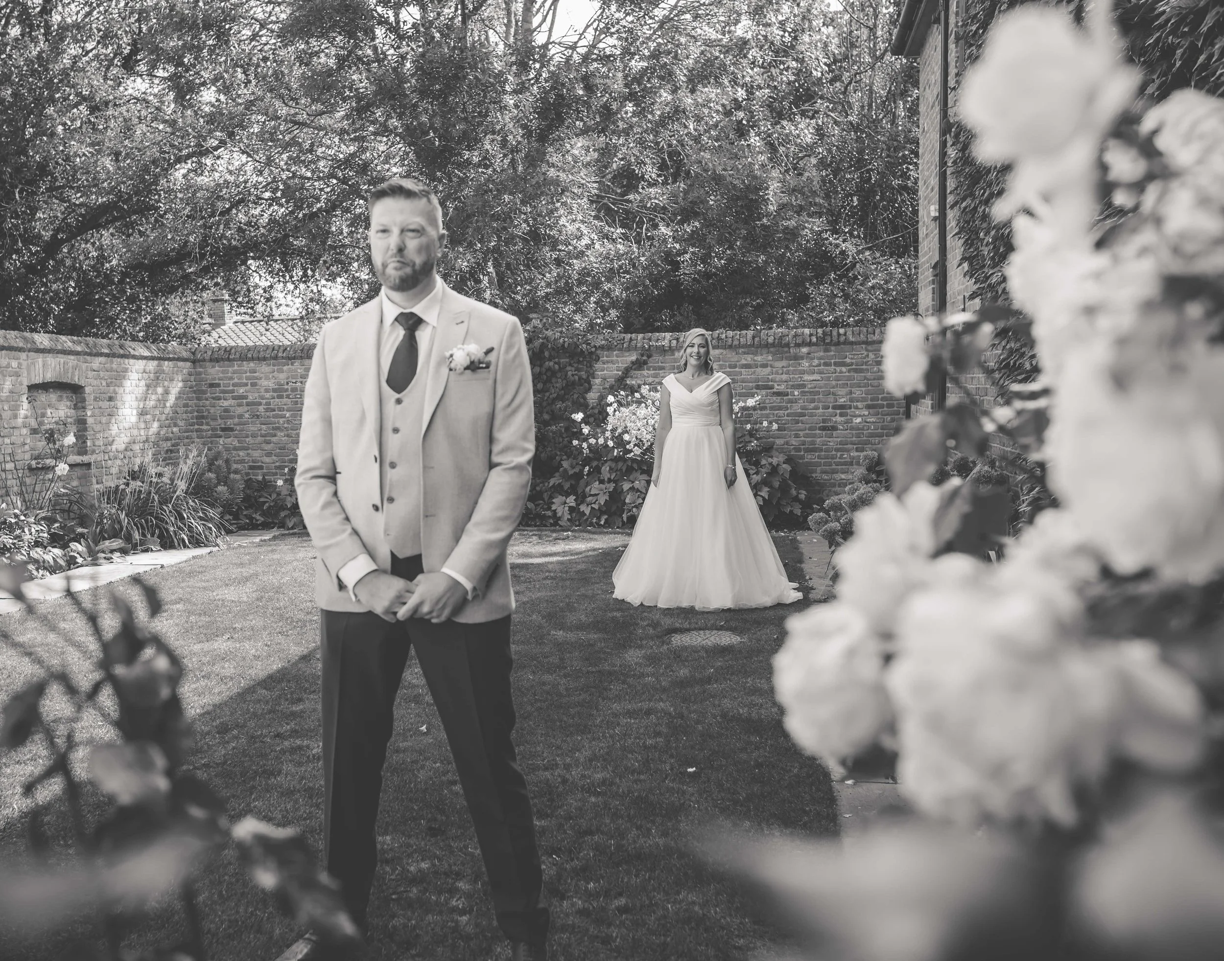 A groom in a light-colored suit with a dark tie and boutonniere stands with a serious expression in a garden. In the background, a bride wearing a white wedding dress and holding a bouquet stands smiling near a brick wall and lush bushes. The photo i