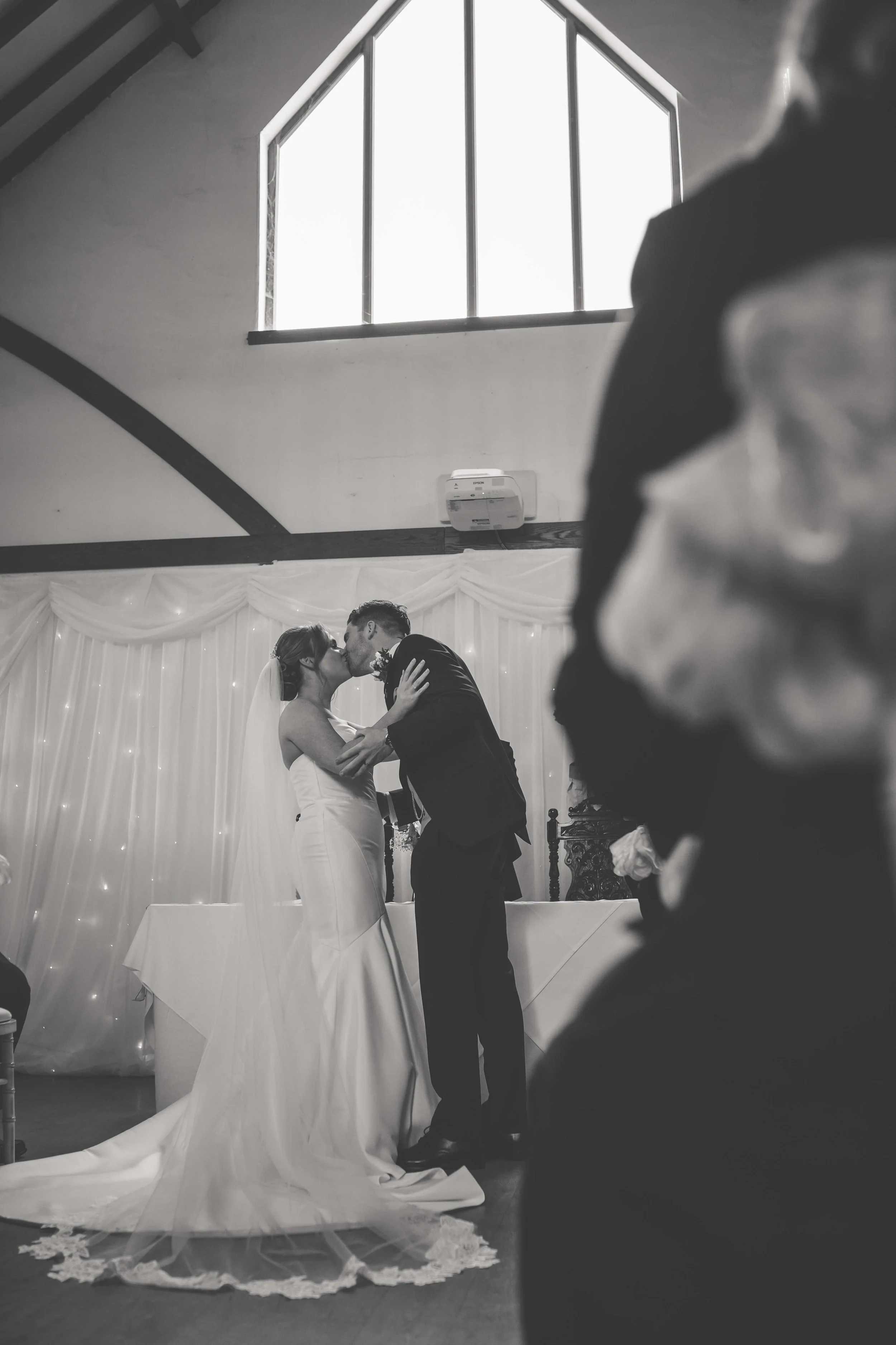 A couple, dressed in wedding attire, sharing a kiss during their ceremony, with a wedding officiant visible in the foreground and a decorated backdrop in the background.