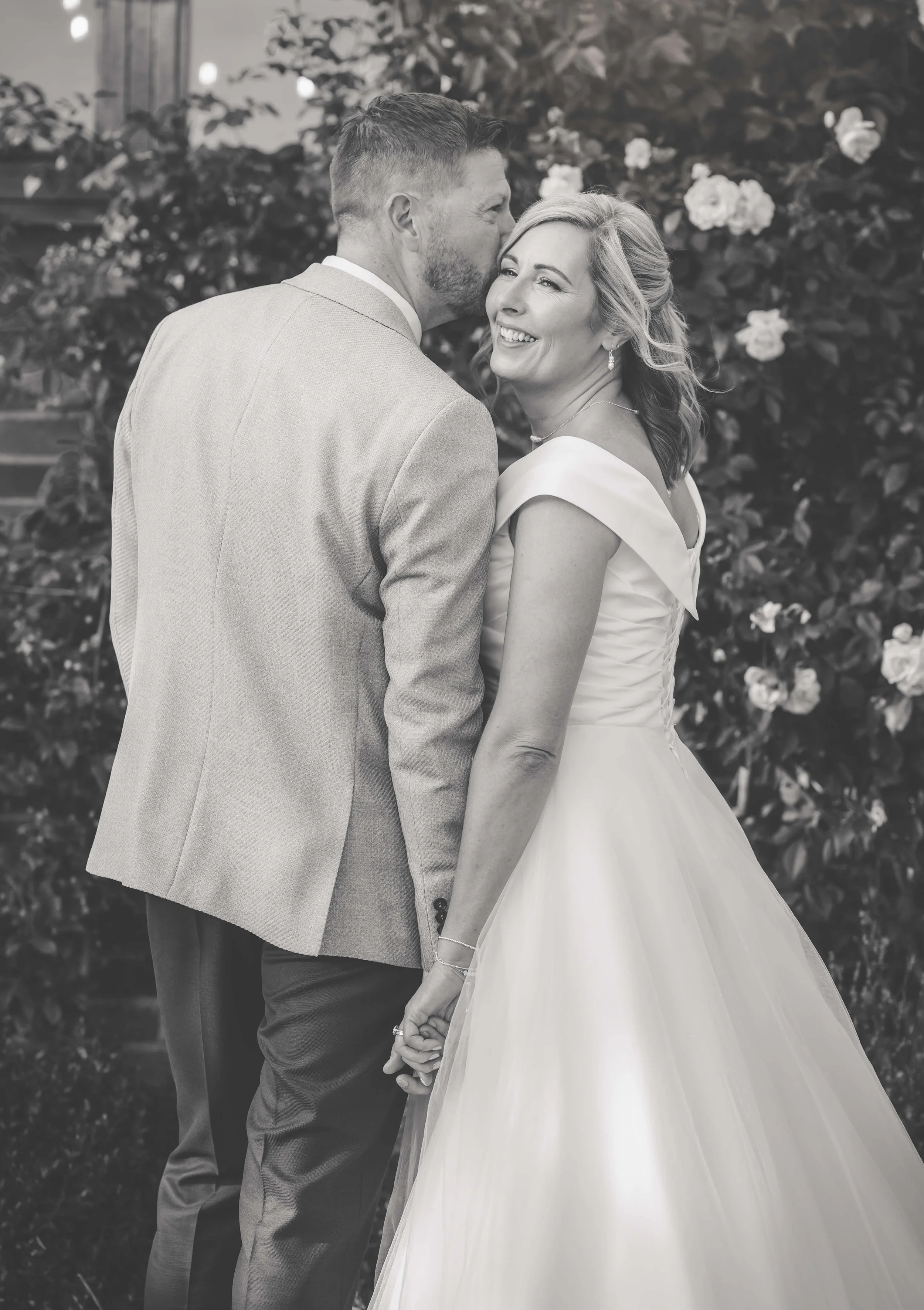 A couple in wedding attire holding hands and smiling, with a background of blooming flowers and greenery.