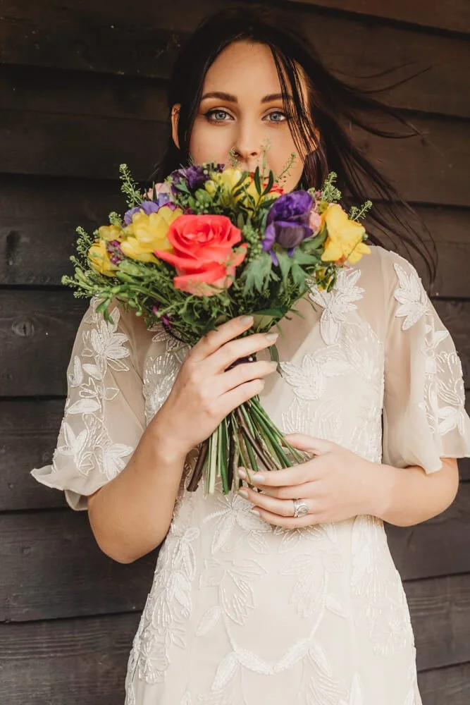 Woman in a white embroidered dress holding a colorful bouquet of flowers in front of her face.