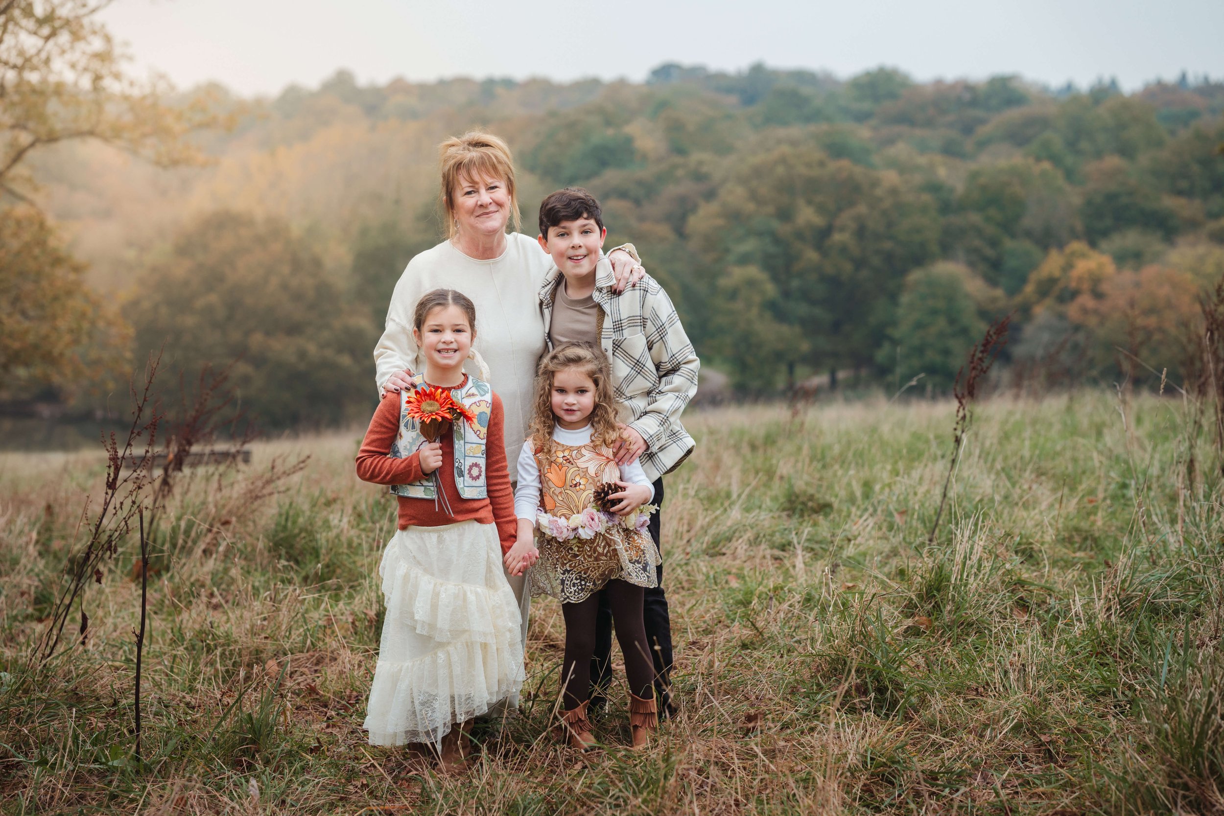 A grandmother with three young children standing in a grassy field with trees in the background during fall.