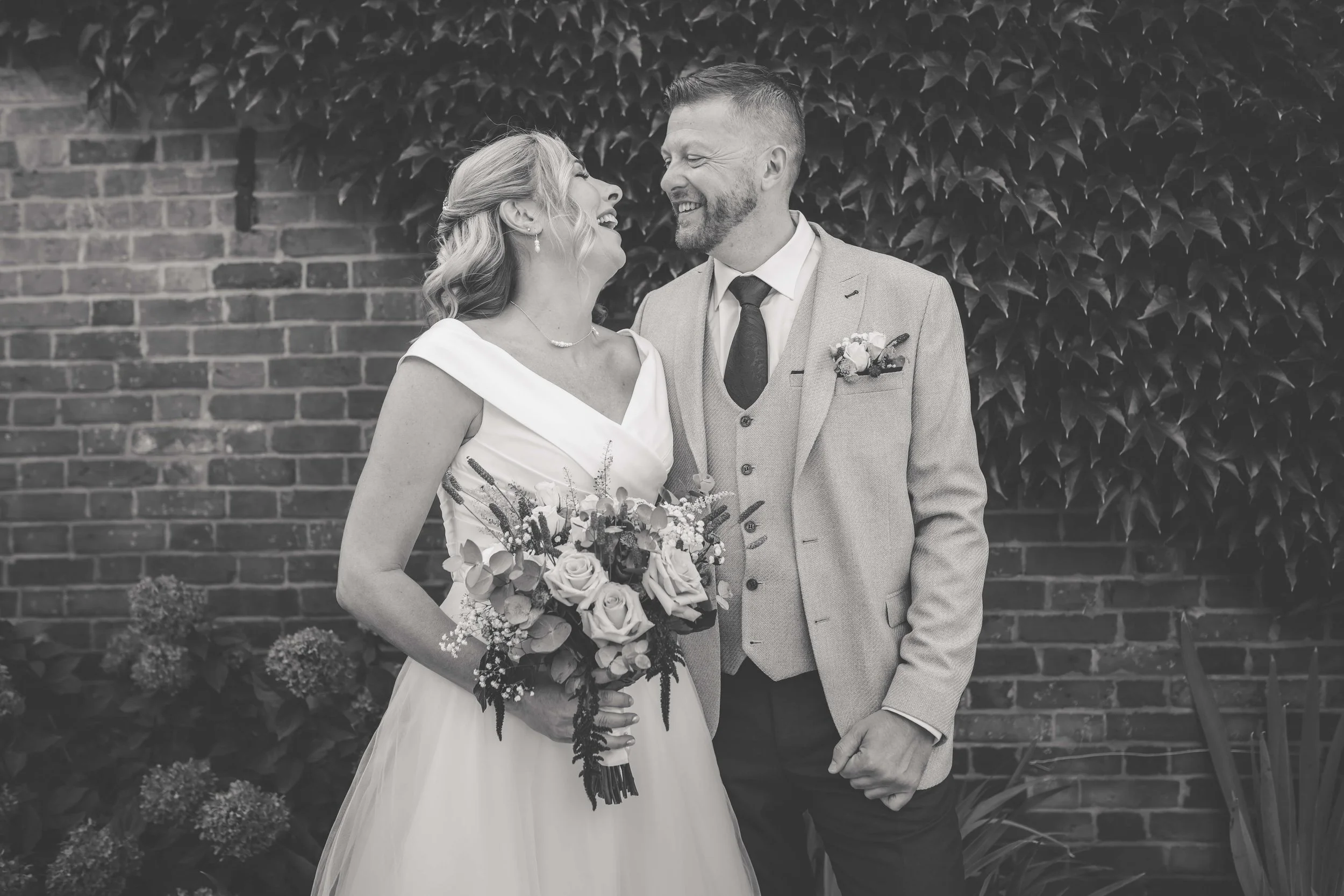 Black and white photo of a bride and groom smiling and looking at each other, standing outdoors in front of a brick wall and leafy plants. The bride is holding a bouquet of flowers.
