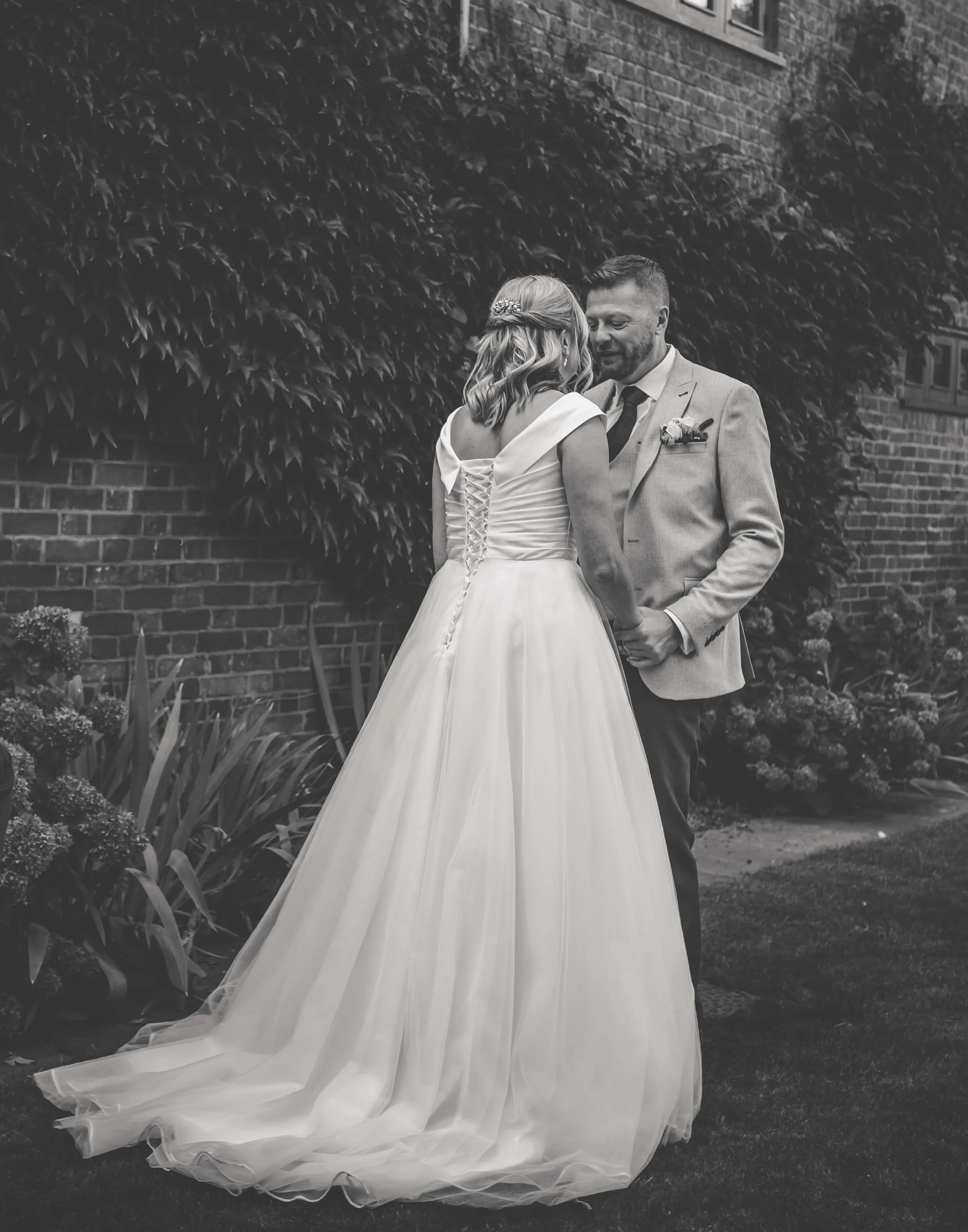 Black and white photo of a bride and groom holding hands and sharing a moment in a garden with a brick wall in the background.