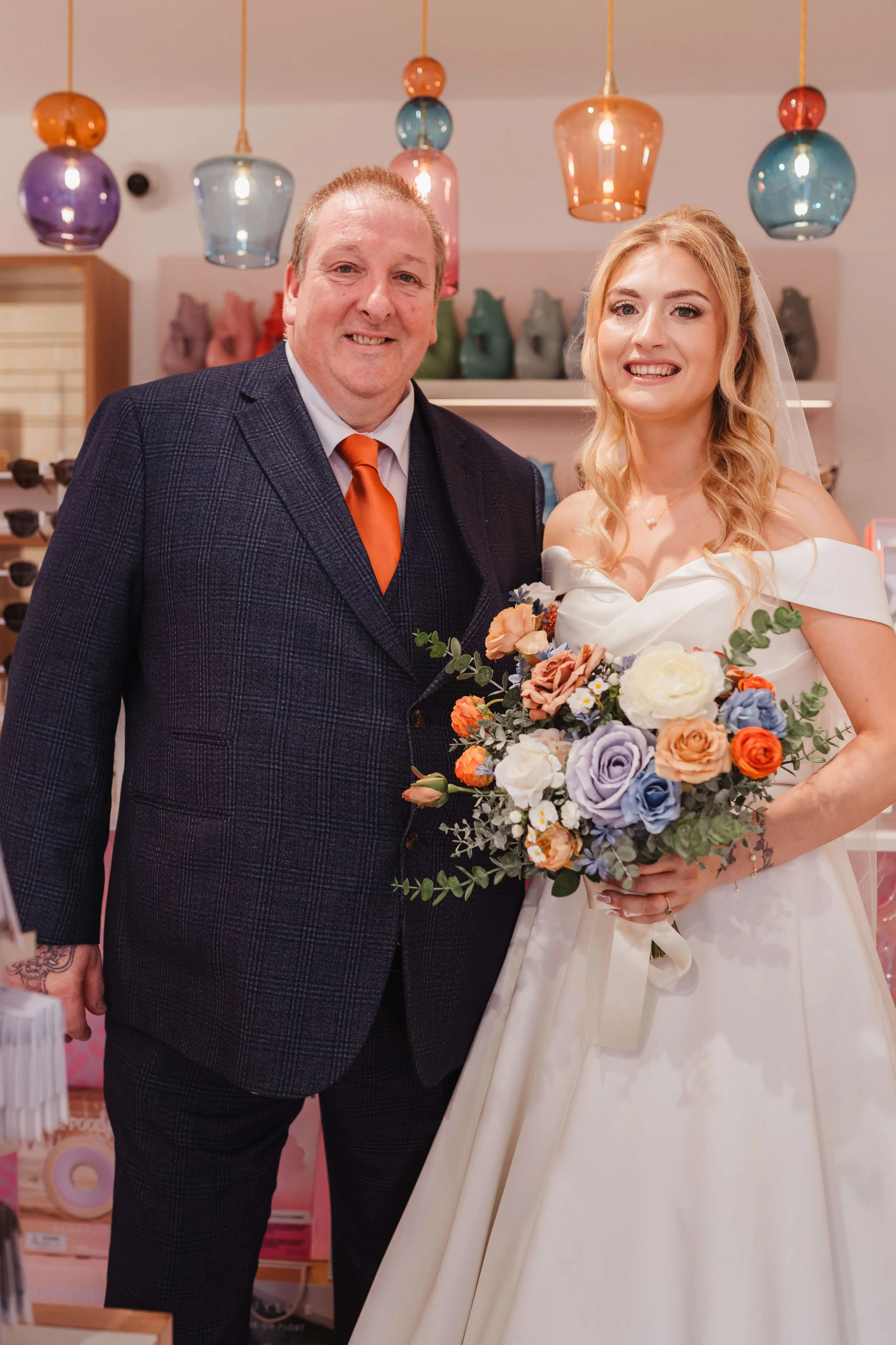 On a wedding day, a smiling bride in a white wedding dress holds a colorful bouquet, standing next to a man in a dark plaid suit with an orange tie, in a store with decorative lighting and colorful ceramics in the background.