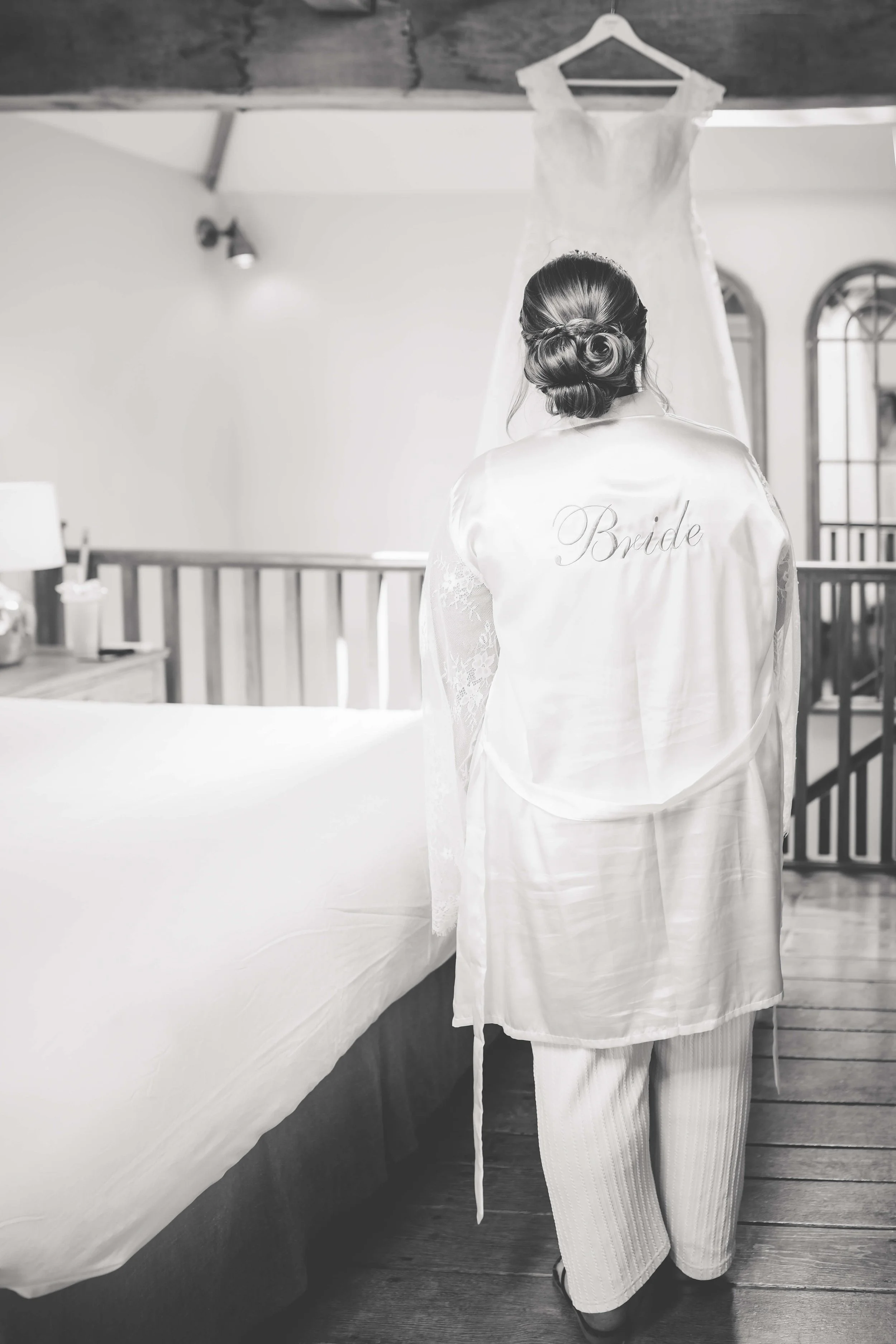 A bride wearing a silk robe with the word 'Bride' embroidered on the back, looking at her wedding dress hanging from the ceiling in a room with wooden floors and a railing