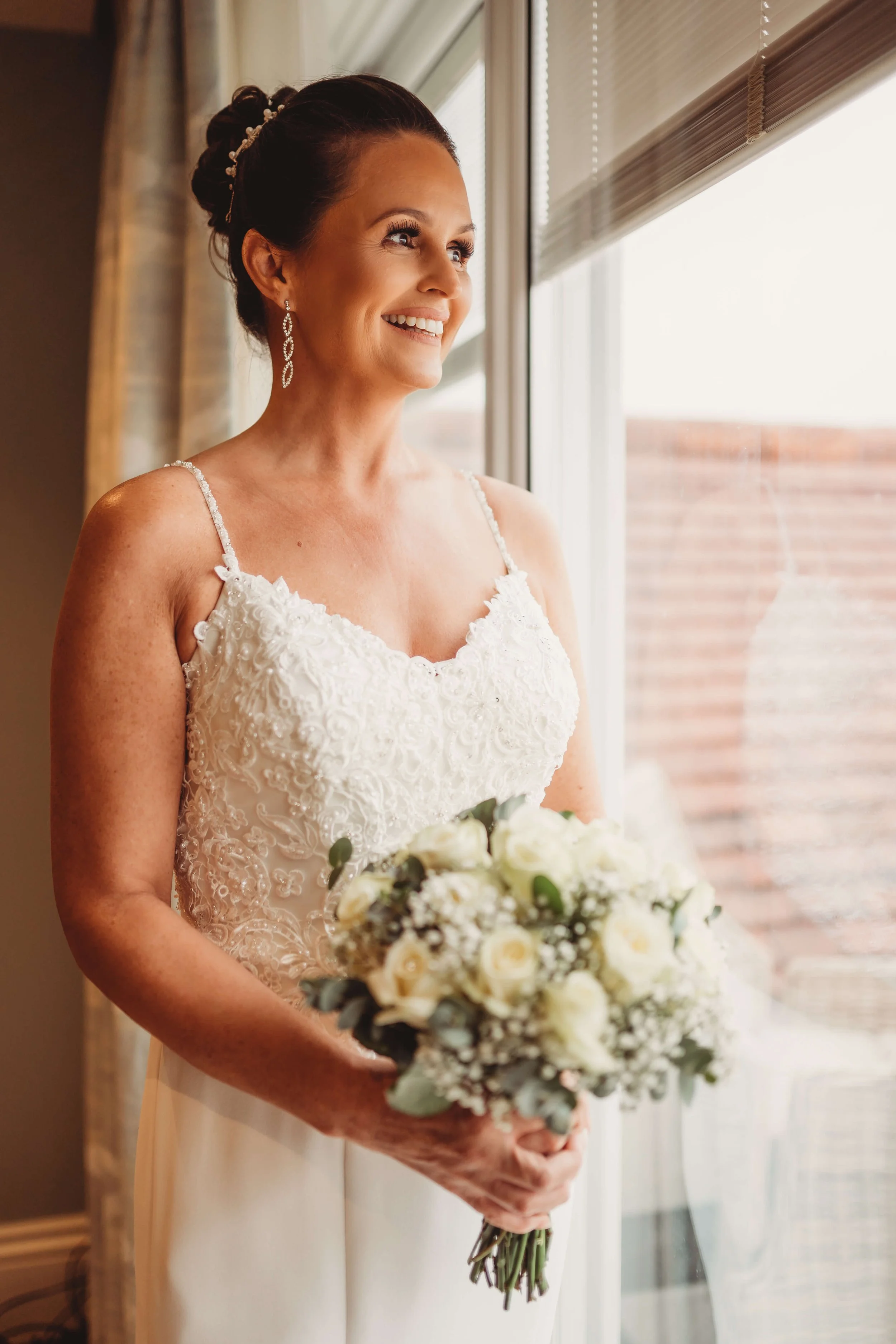 A woman in a white wedding dress holding a bouquet of white roses and greenery, standing near a window with blinds, smiling and looking outside.