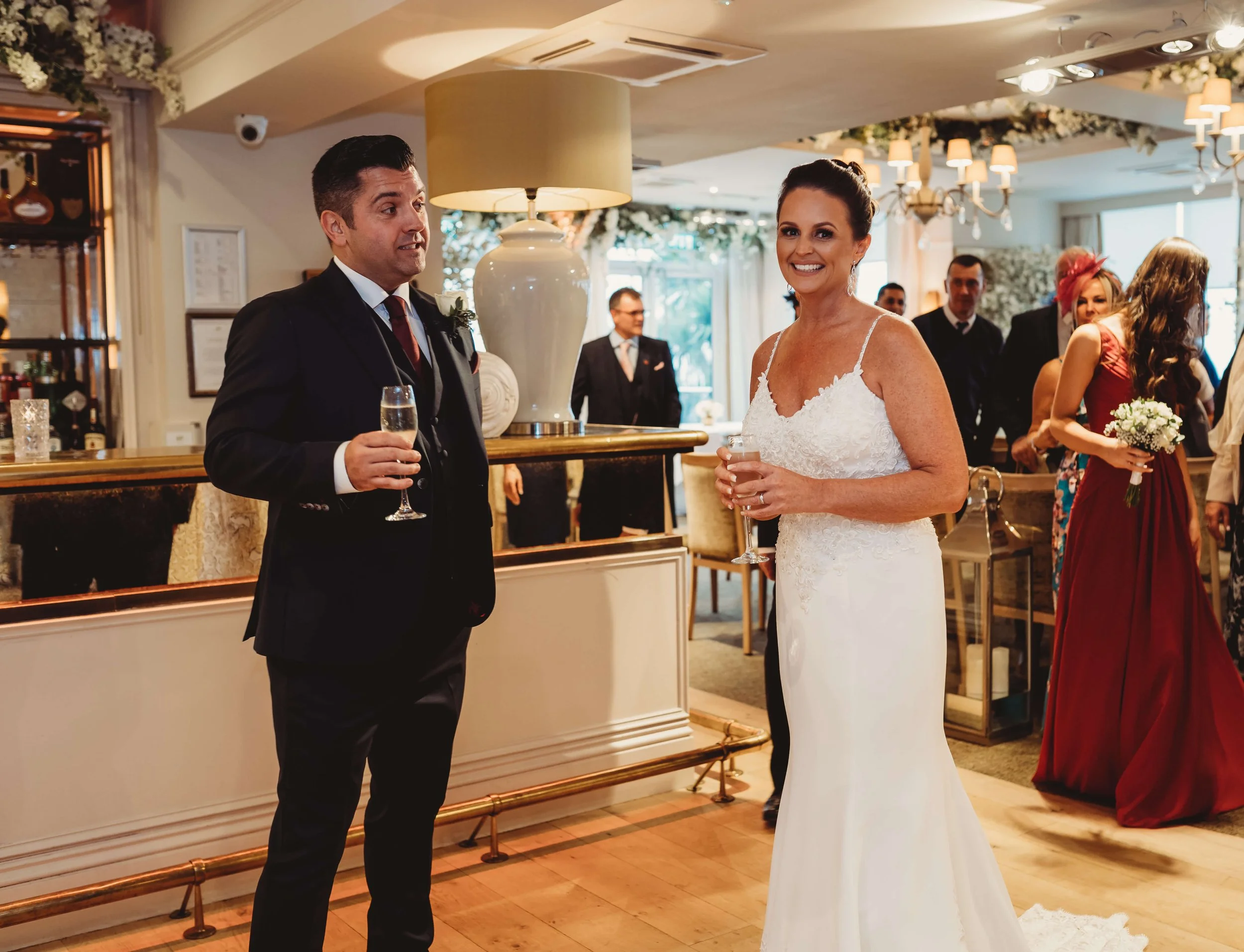 Bride and groom at wedding reception talking and holding champagne glasses in a decorated indoor venue with guests in the background.