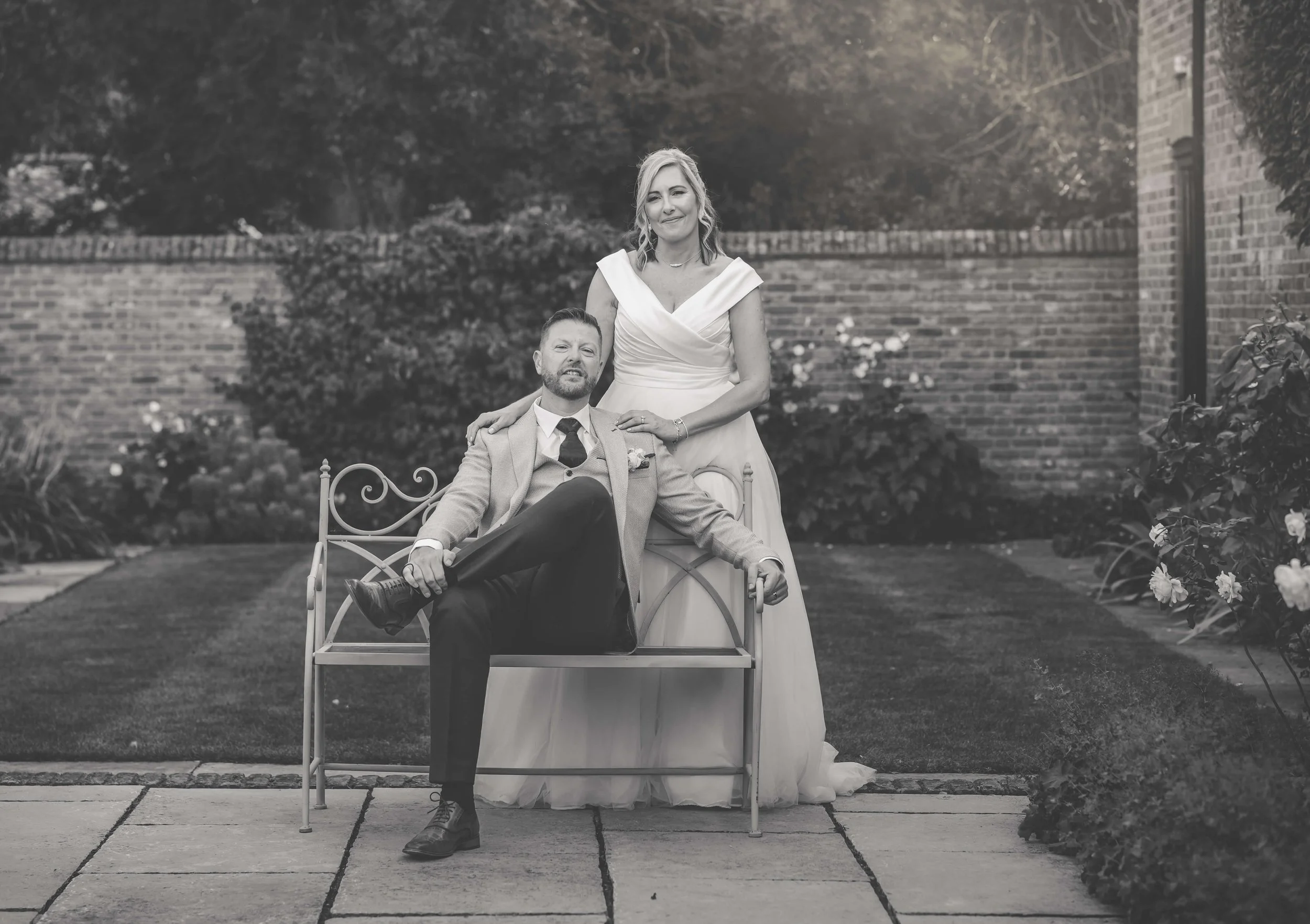 Black and white photo of a couple outdoors. The woman is standing and wearing a wedding dress, and the man is sitting on a bench, wearing a suit. The woman has her hand on the man's shoulder, and they are posing for the camera.