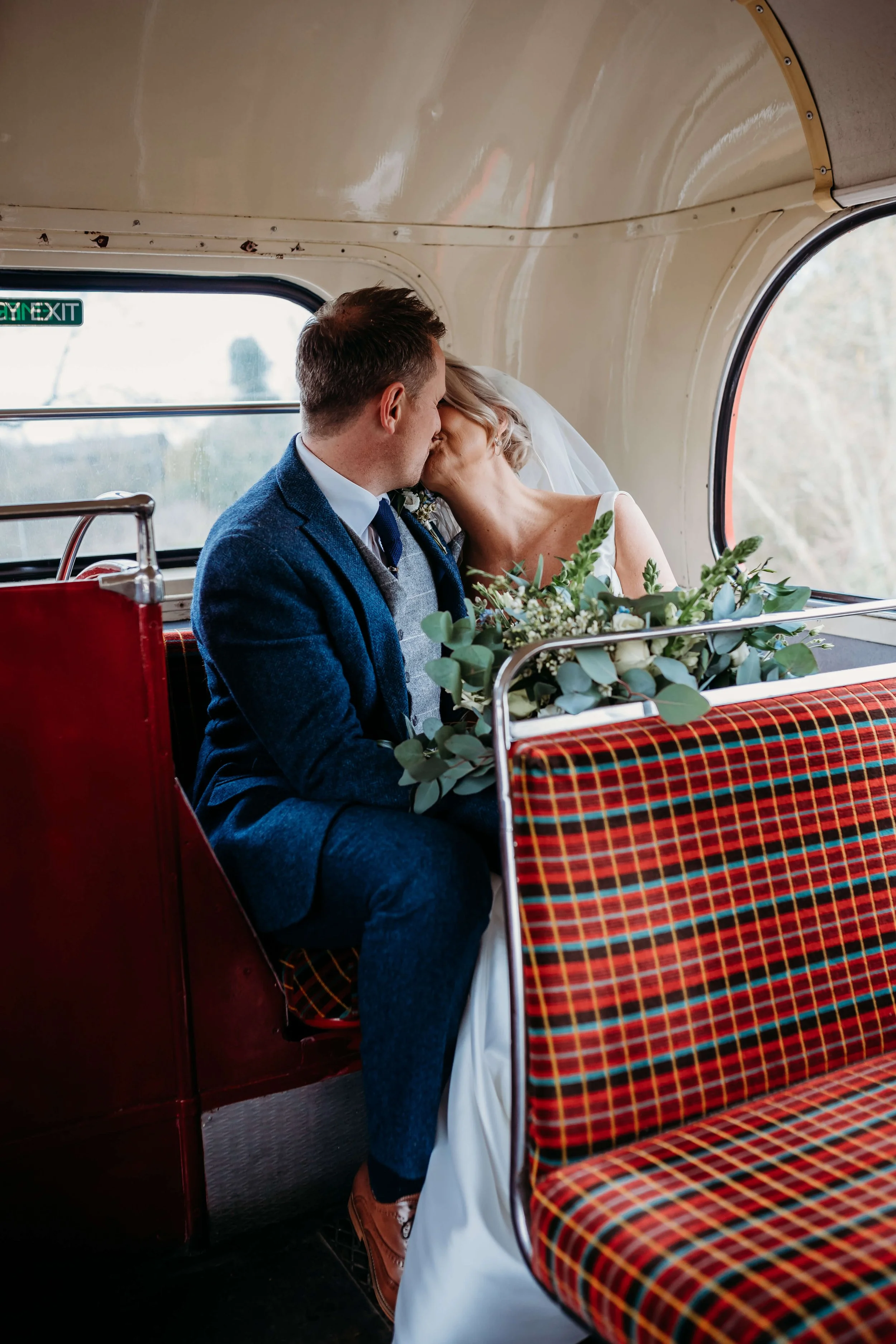 A bride and groom sharing a kiss on a bus, with the bride holding a bouquet of green foliage.