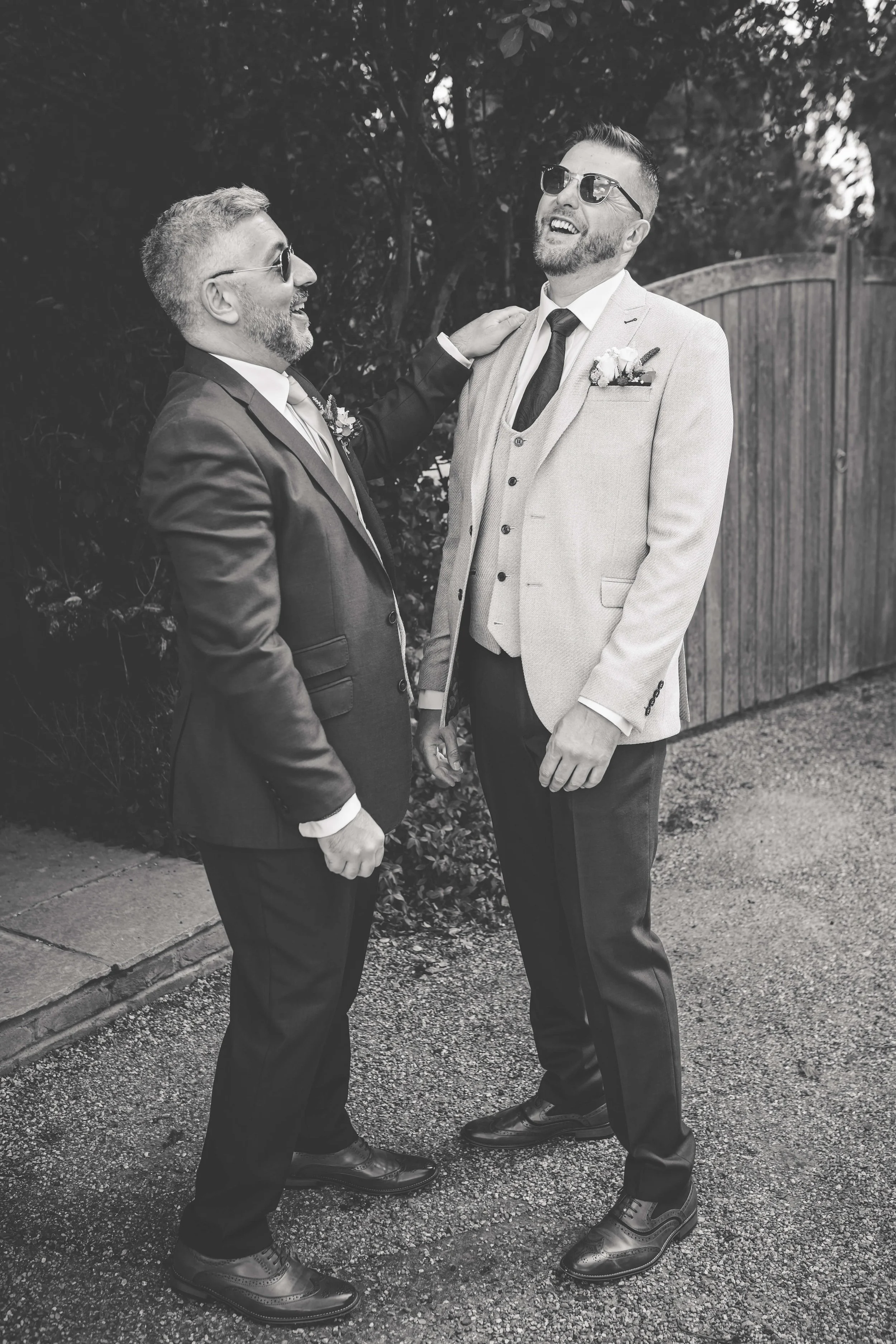 Two men dressed in suits, one in a light-colored blazer and the other in a darker suit, laughing and holding hands outdoors.