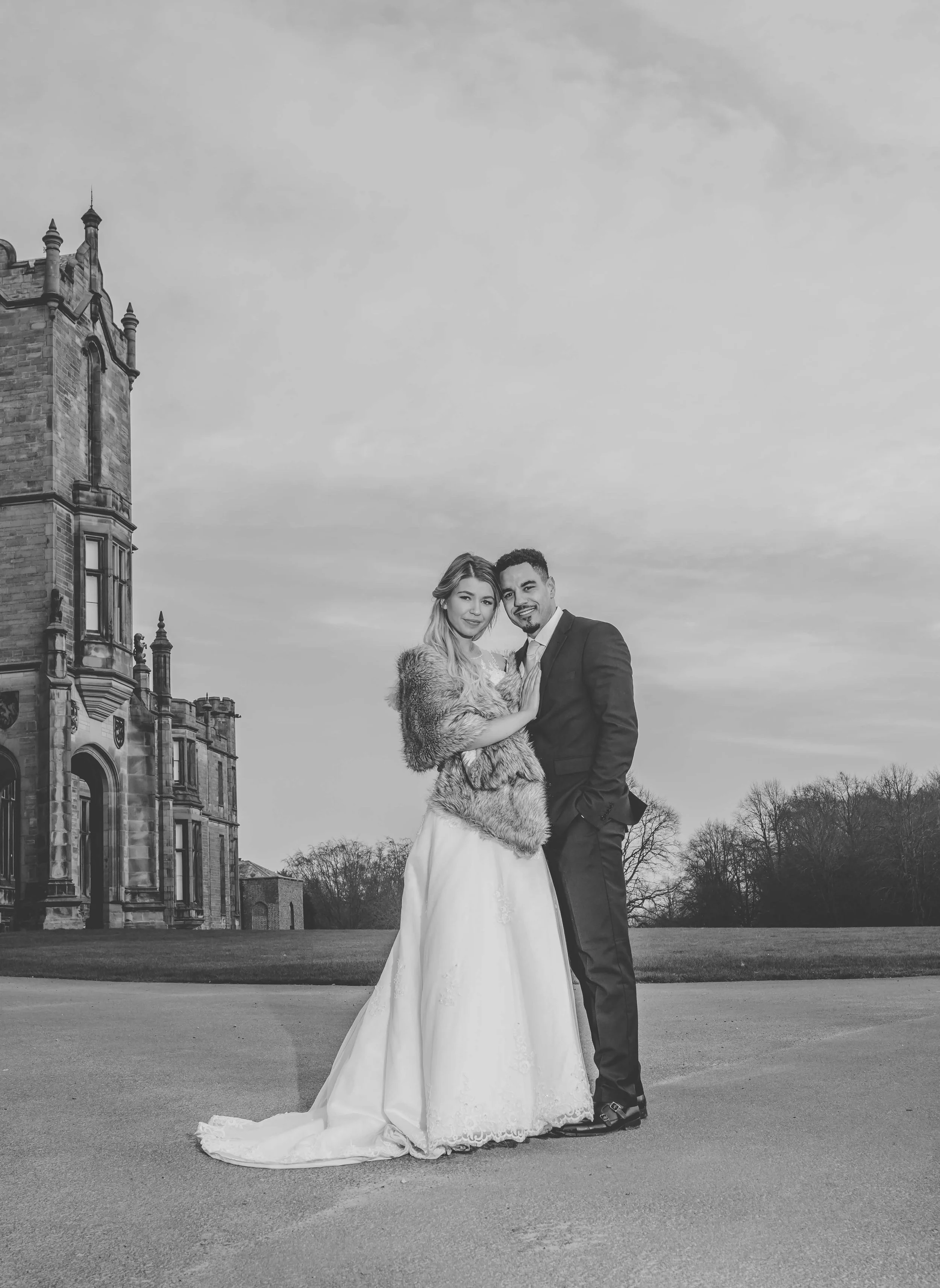 Black and white photo of a bride and groom standing outside in front of a large historic building with a lawn and trees in the background. The bride is wearing a wedding gown and a fur stole, and the groom is in a formal suit. They are smiling and em