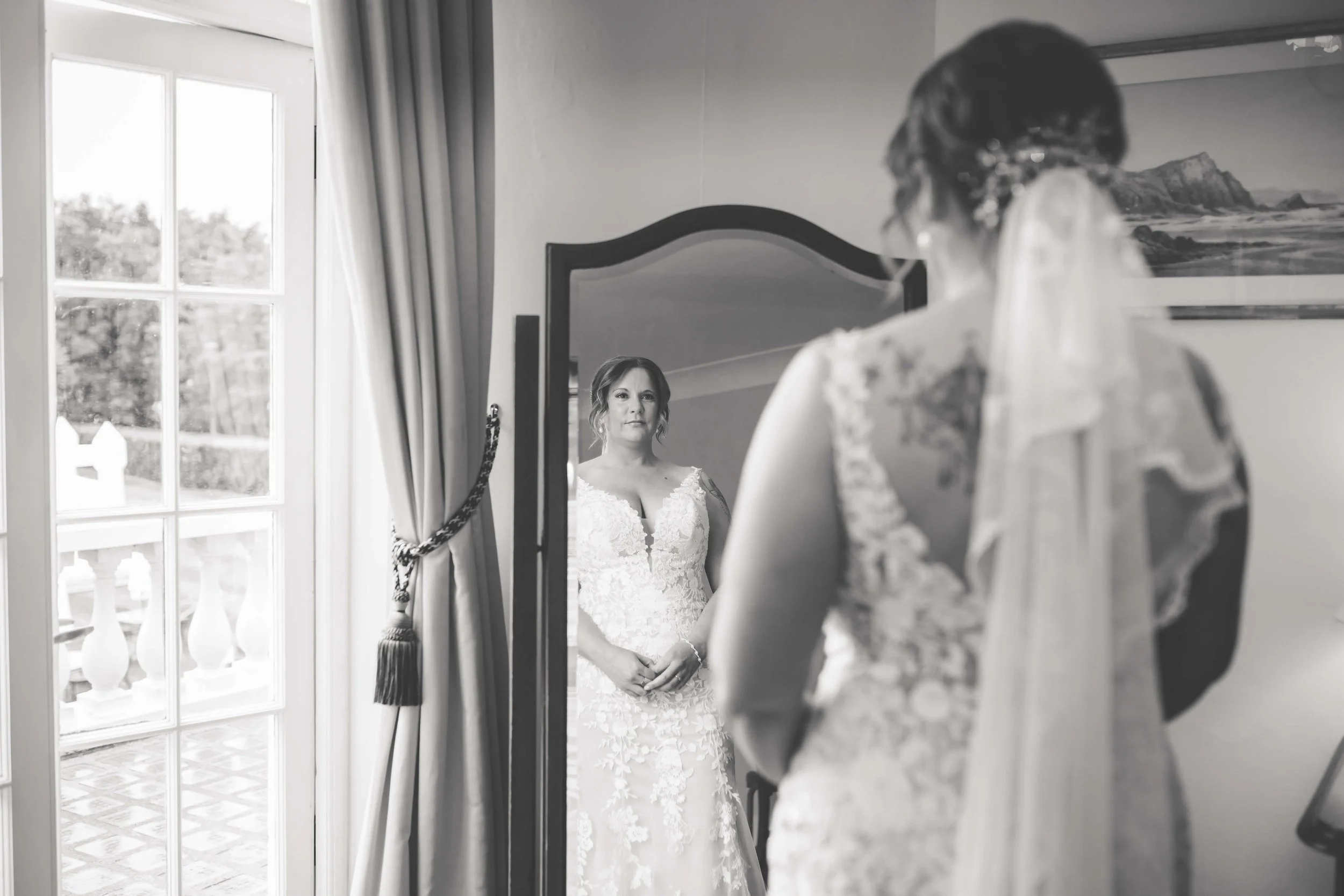 A bride standing in front of a mirror, looking at her reflection in a room with large window and curtains.