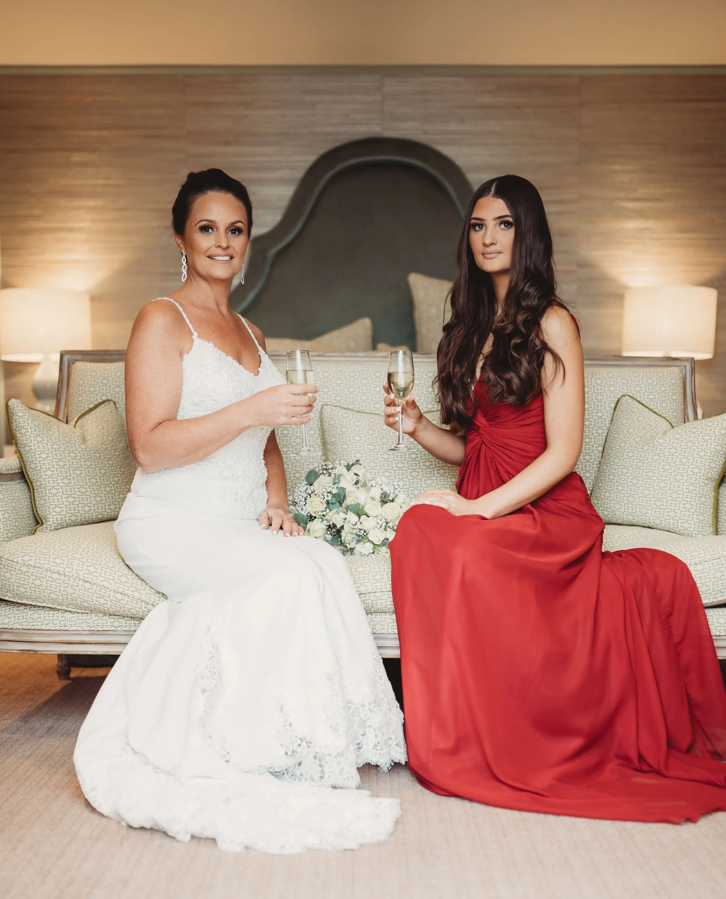 Two women in elegant dresses sitting on a beige sofa, holding glasses of champagne, with a bridal bouquet on the sofa between them, in a warmly lit room.