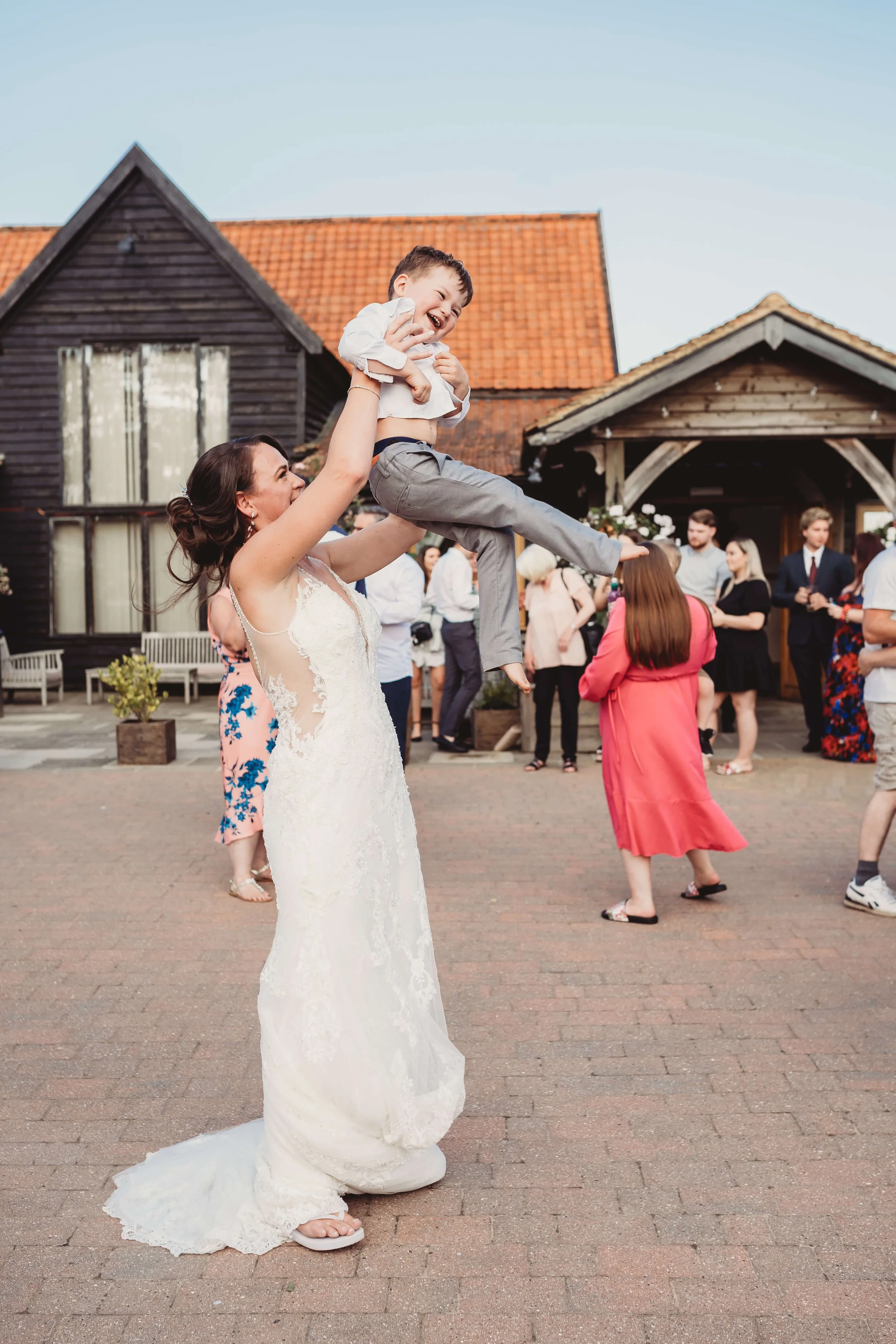 A woman in a wedding dress lifting a young boy in a white shirt and gray pants in the air at an outdoor event, with people mingling in the background.