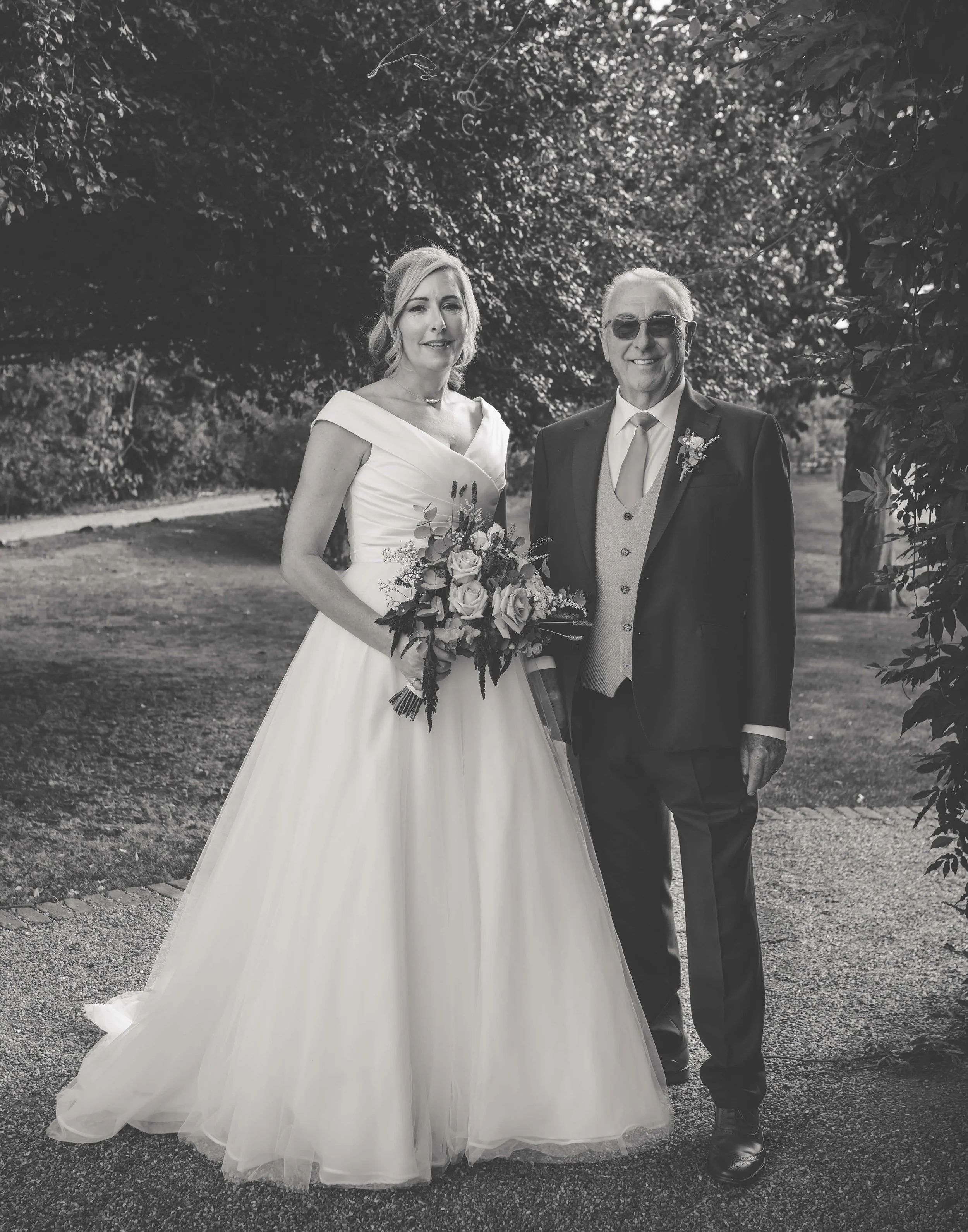 Black and white photo of a bride in a wedding gown holding a bouquet of flowers standing beside an older man in a suit outdoors.