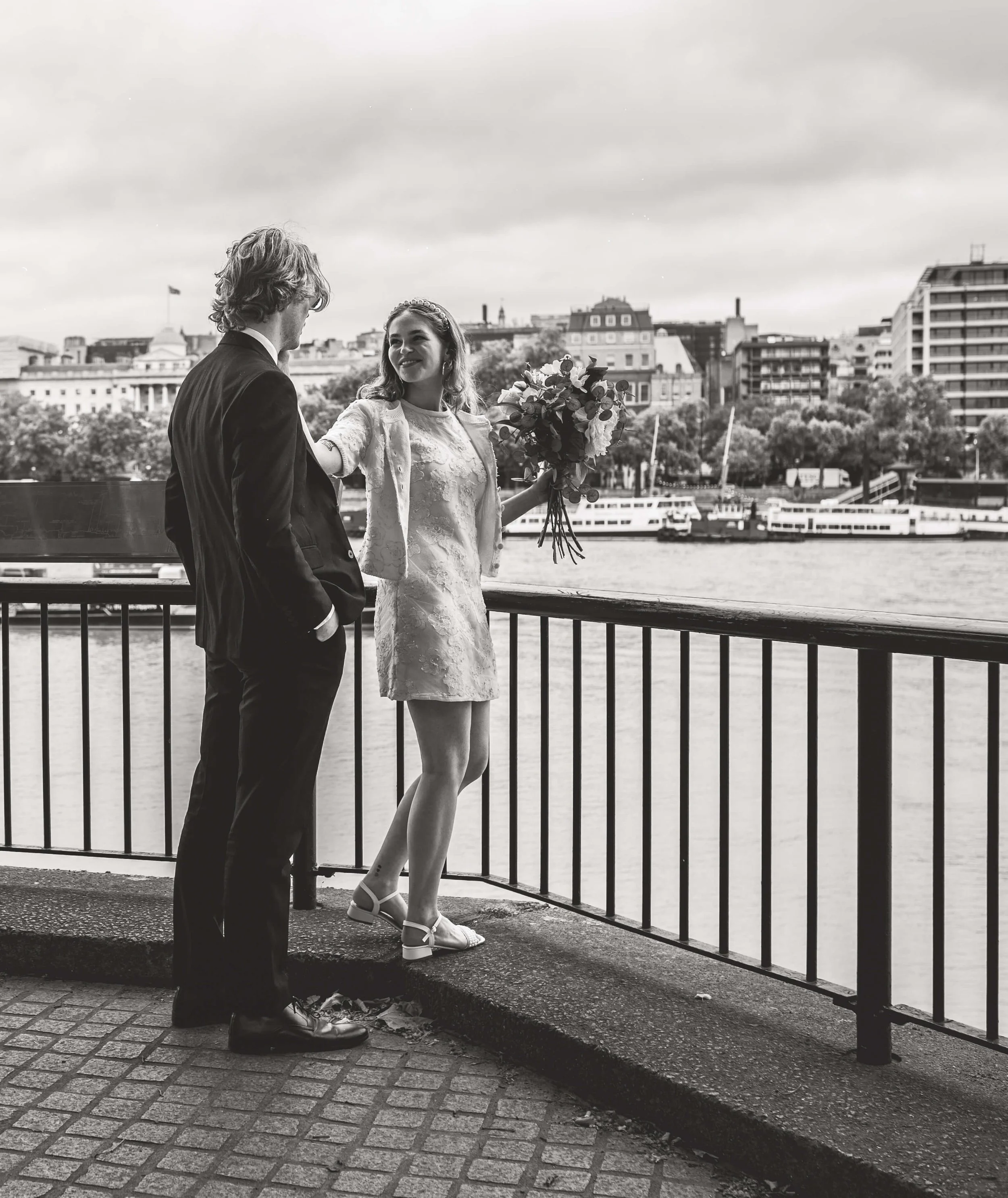 A black-and-white photo of a man and woman on a riverside promenade. The woman, holding a bouquet of flowers, is smiling and reaching out to the man. The man is dressed in a suit and has his hands in his pockets. In the background, there are building