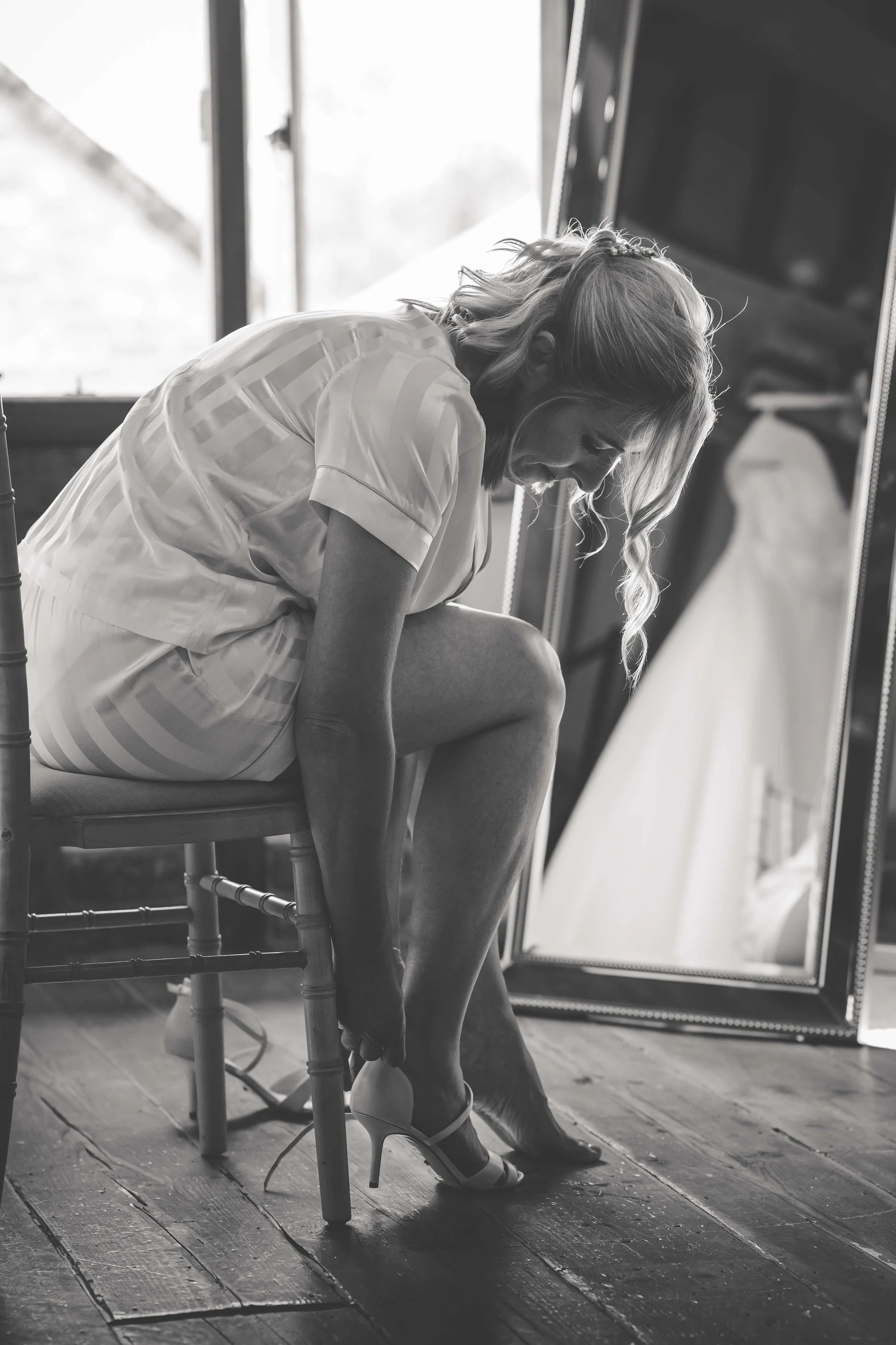 A woman in a short dress putting on high heels, sitting on a chair near a mirror.
