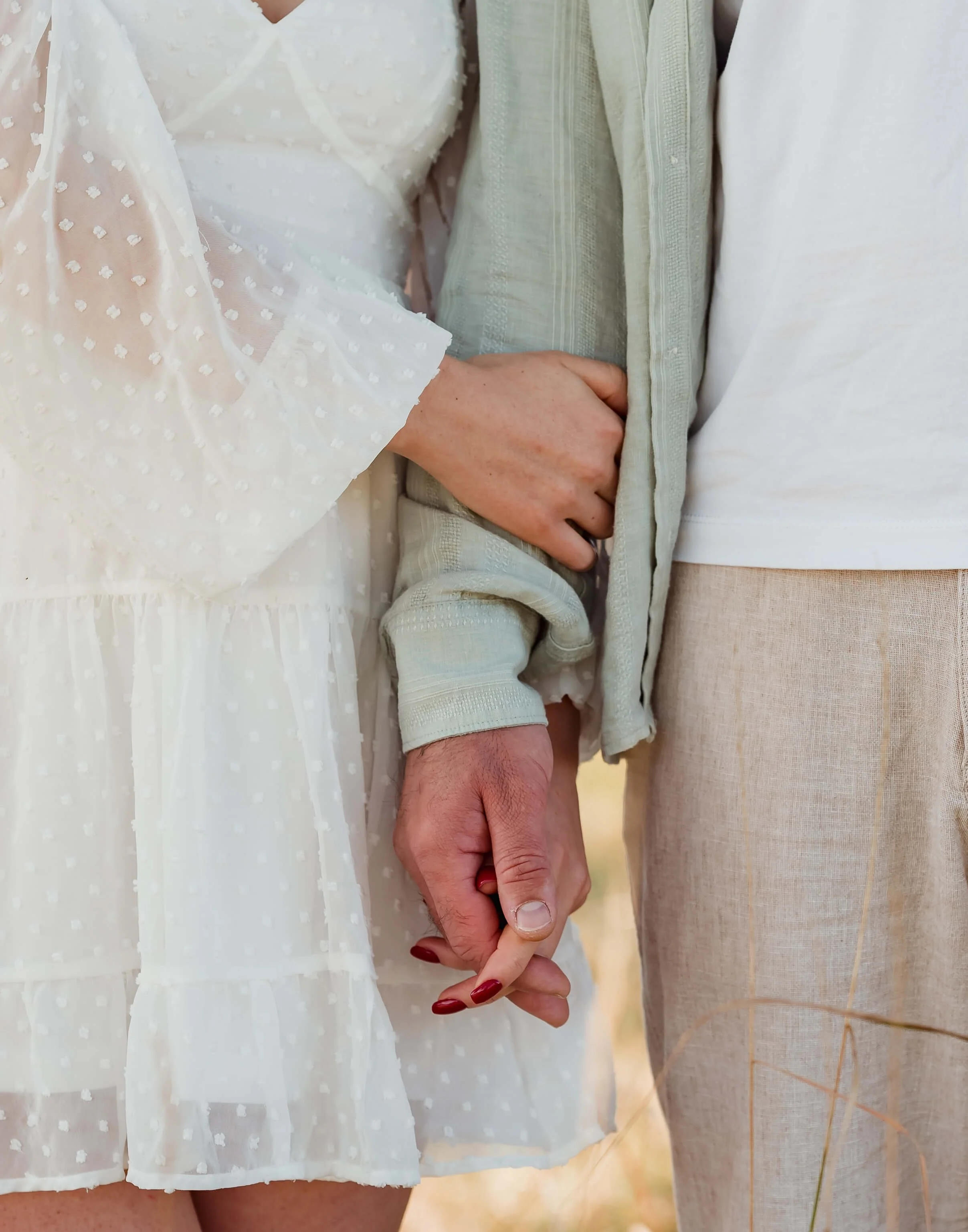 A woman and man holding hands. She is wearing a white dress with sheer fabric and small dot patterns. He is wearing a light-colored jacket and beige pants.