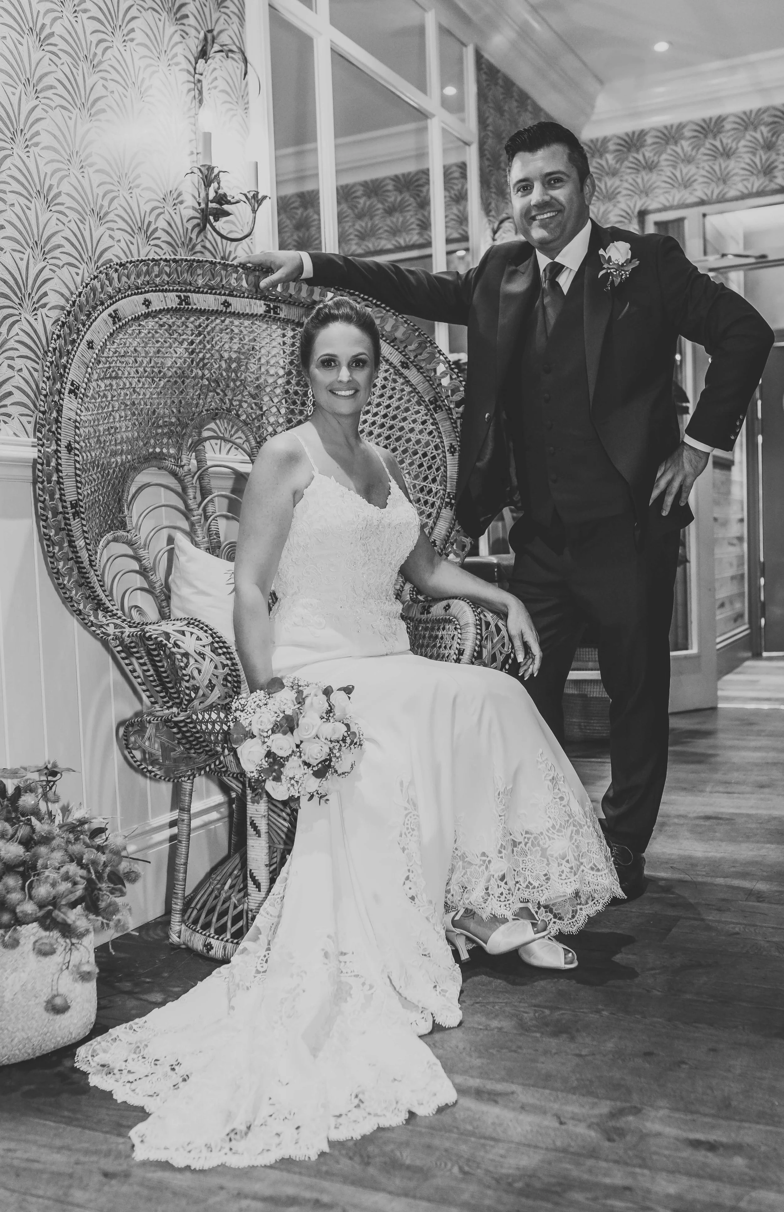 A black and white photo of a bride and groom indoors, with the groom standing behind the bride who is seated on a decorative wicker chair, holding a bouquet of flowers. The groom is wearing a dark suit with a flower boutonniere, and the bride is wear