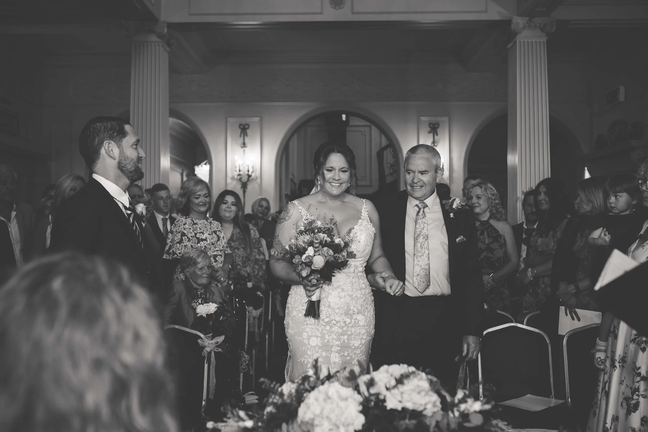 Black and white photograph of a woman in a lace wedding dress being walked down the aisle by an older man, with guests seated and watching in a grand, ornate room.
