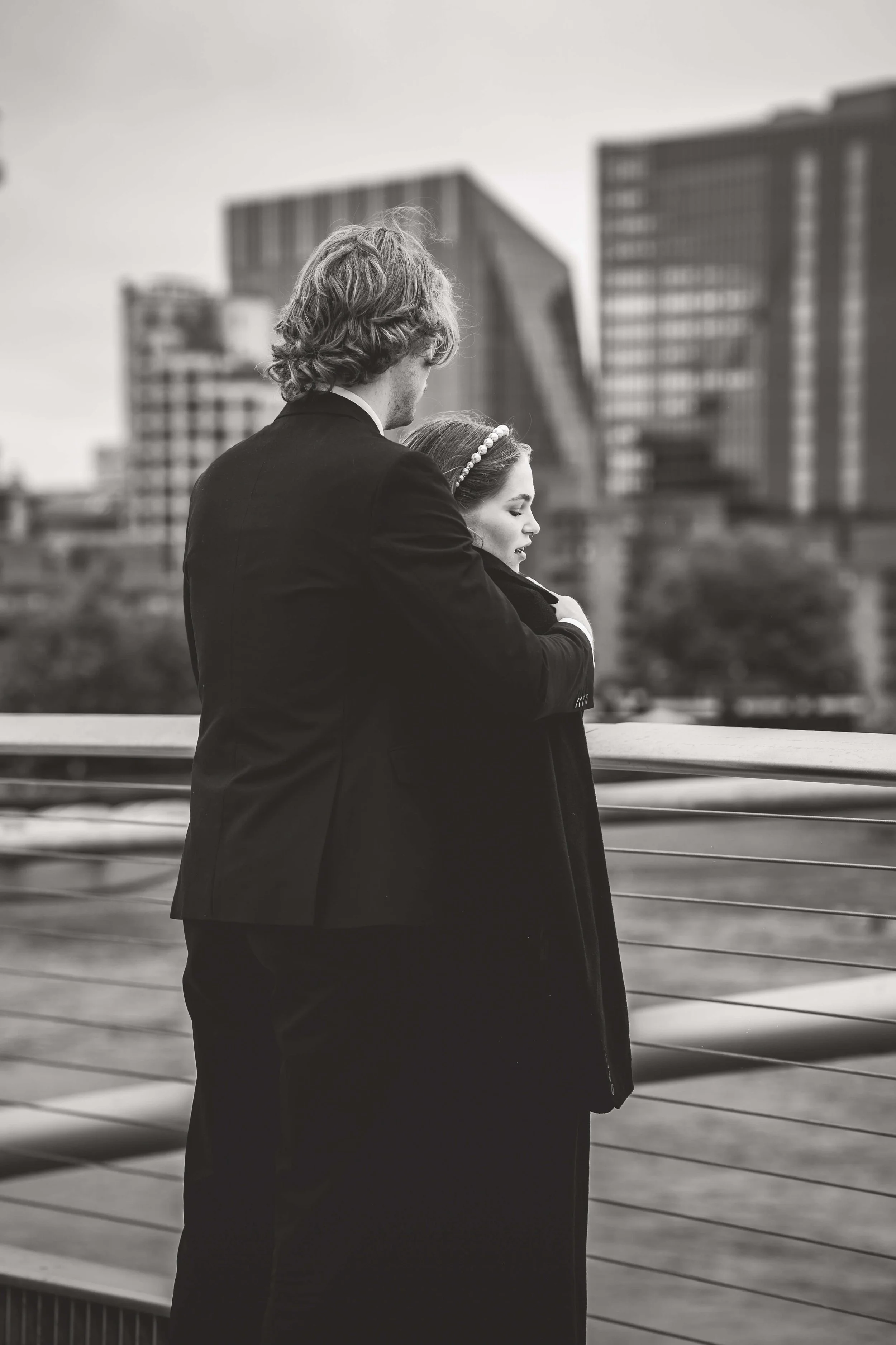 A black and white photo of a man in a suit and a woman with a headband embracing on a bridge with a cityscape in the background.