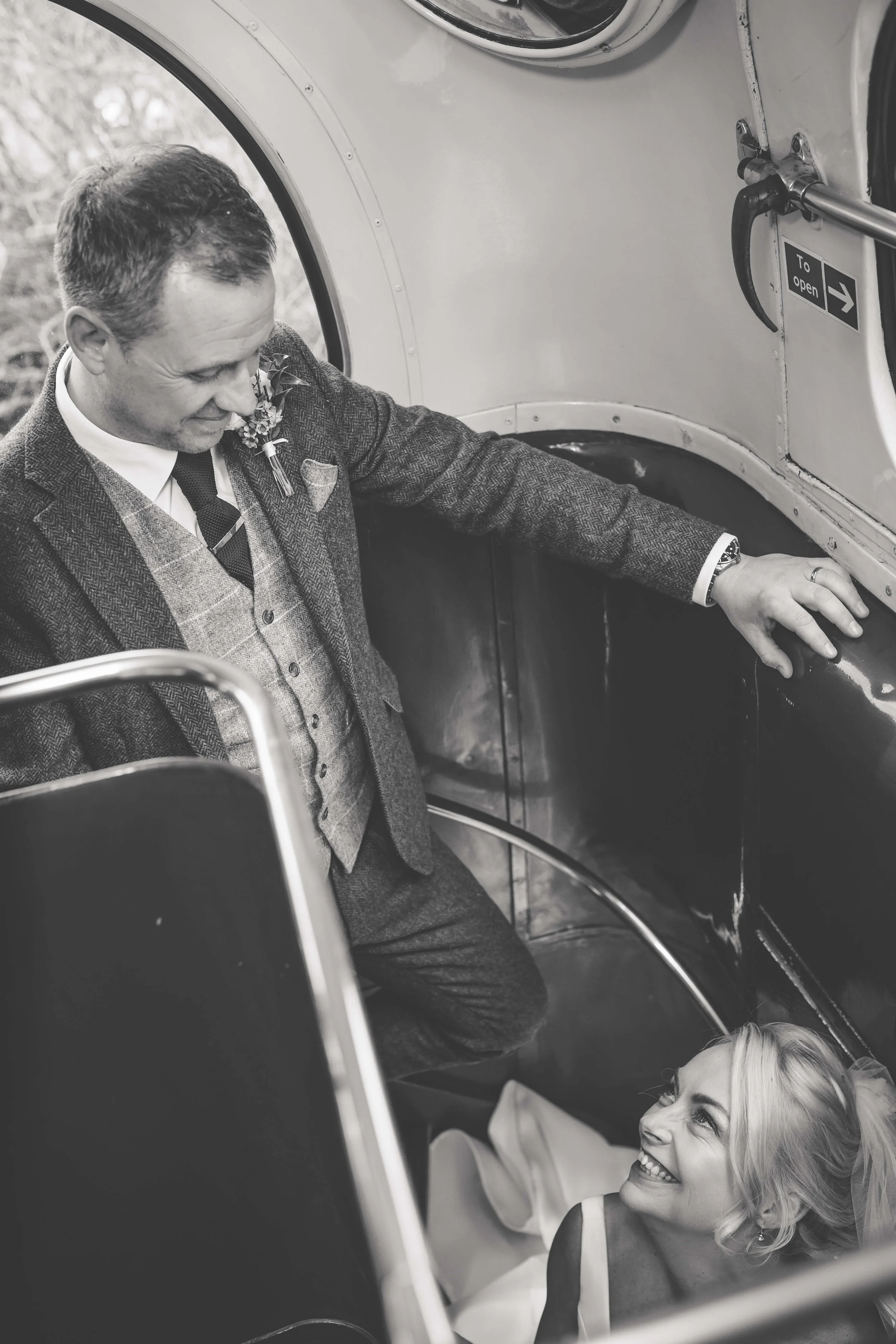 A black-and-white photo of a man in a suit bending down to talk to a woman lying in an emergency exit of a small airplane, both smiling at each other.