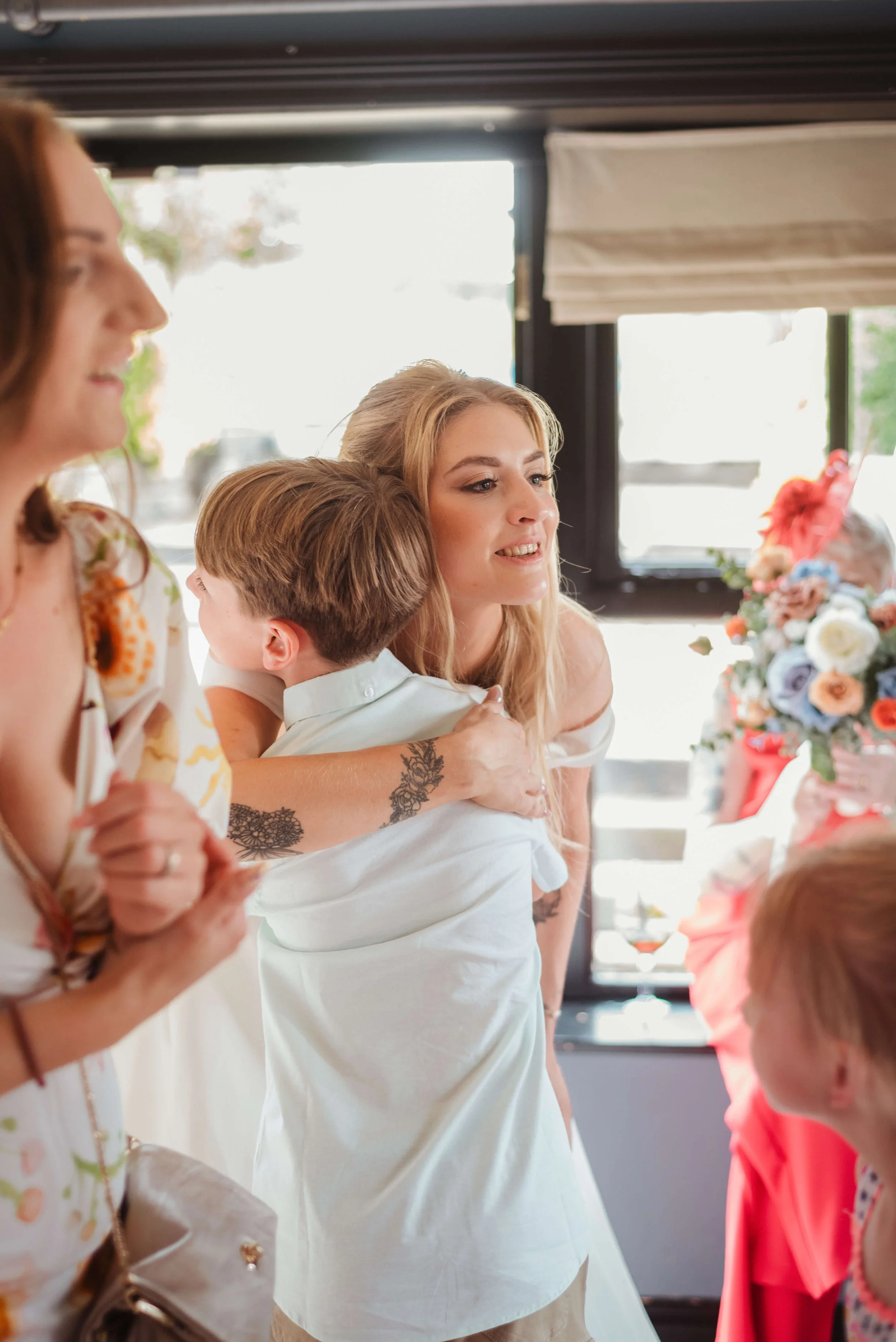 A woman in a white dress hugging a young boy, with other women and children nearby, in a brightly lit room with large windows and floral decorations.