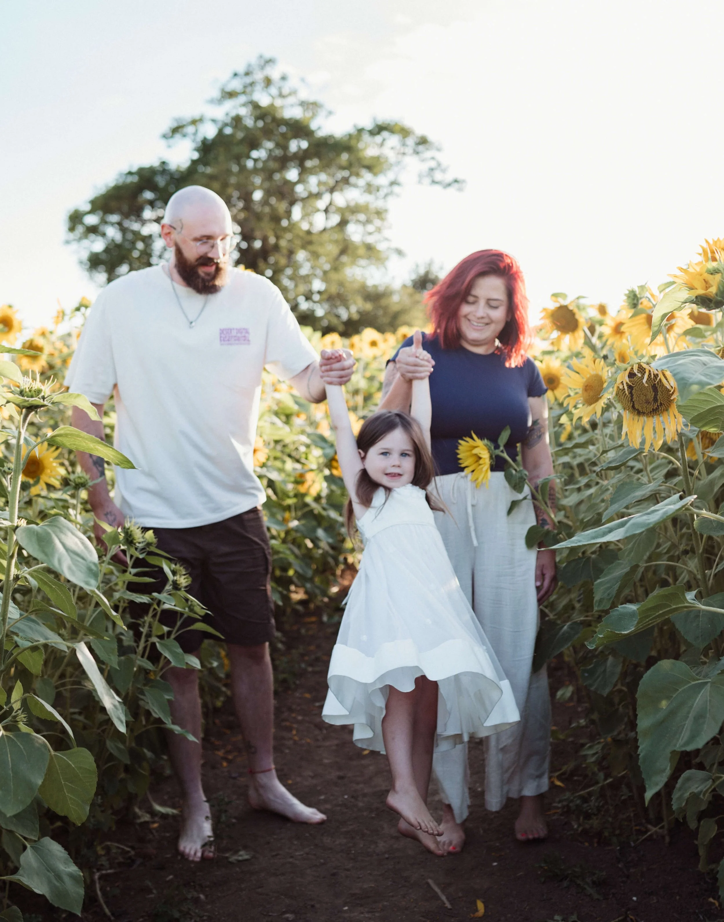 A family of three, a man, a woman, and a young girl, walking through a sunflower field holding hands and smiling.