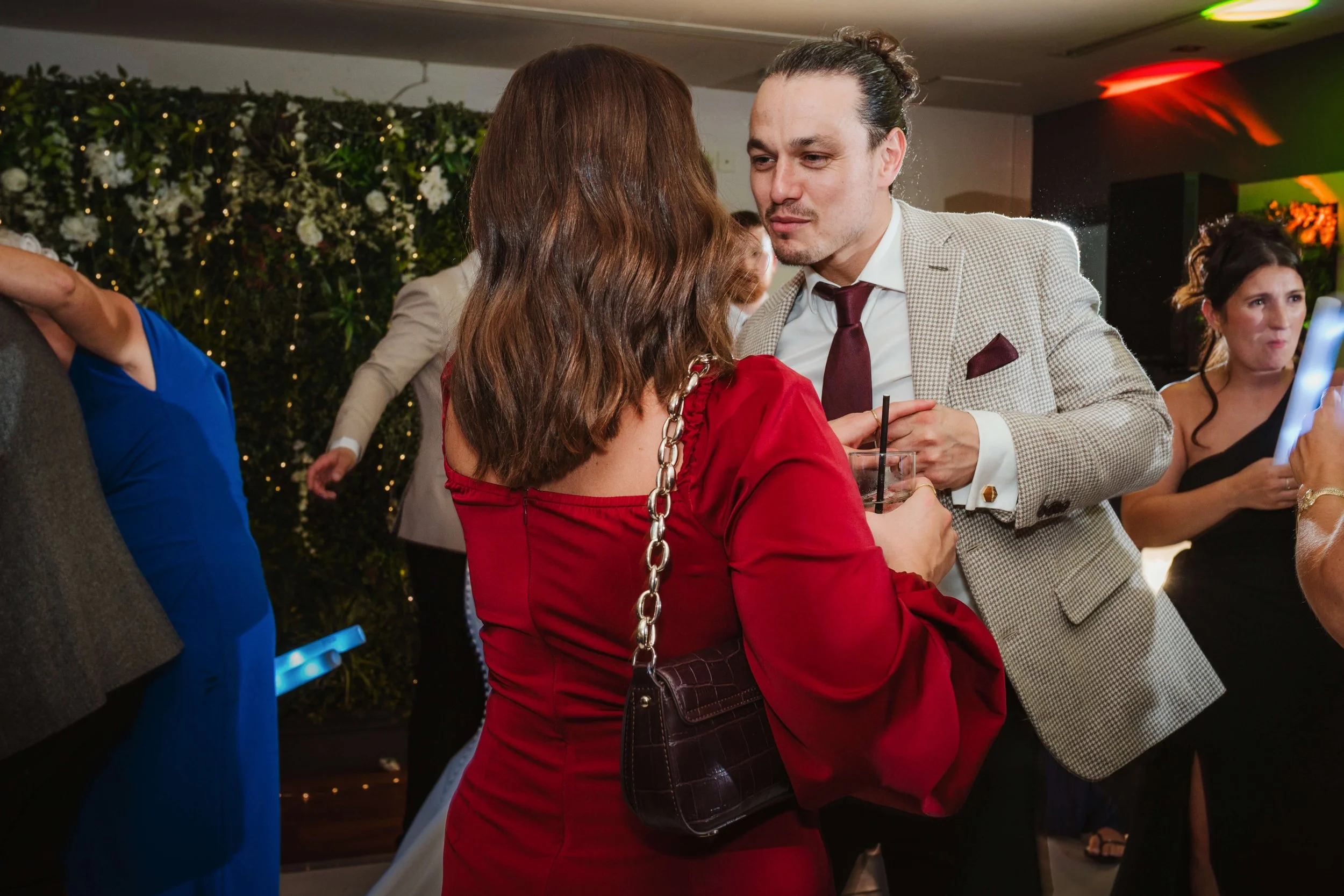 A man and a woman are in conversation at a party, with drinks in their hands. The man is wearing a light-colored checked suit and tie, and the woman is dressed in a red dress with a chain strap purse. Other people are seen in the background at the ev