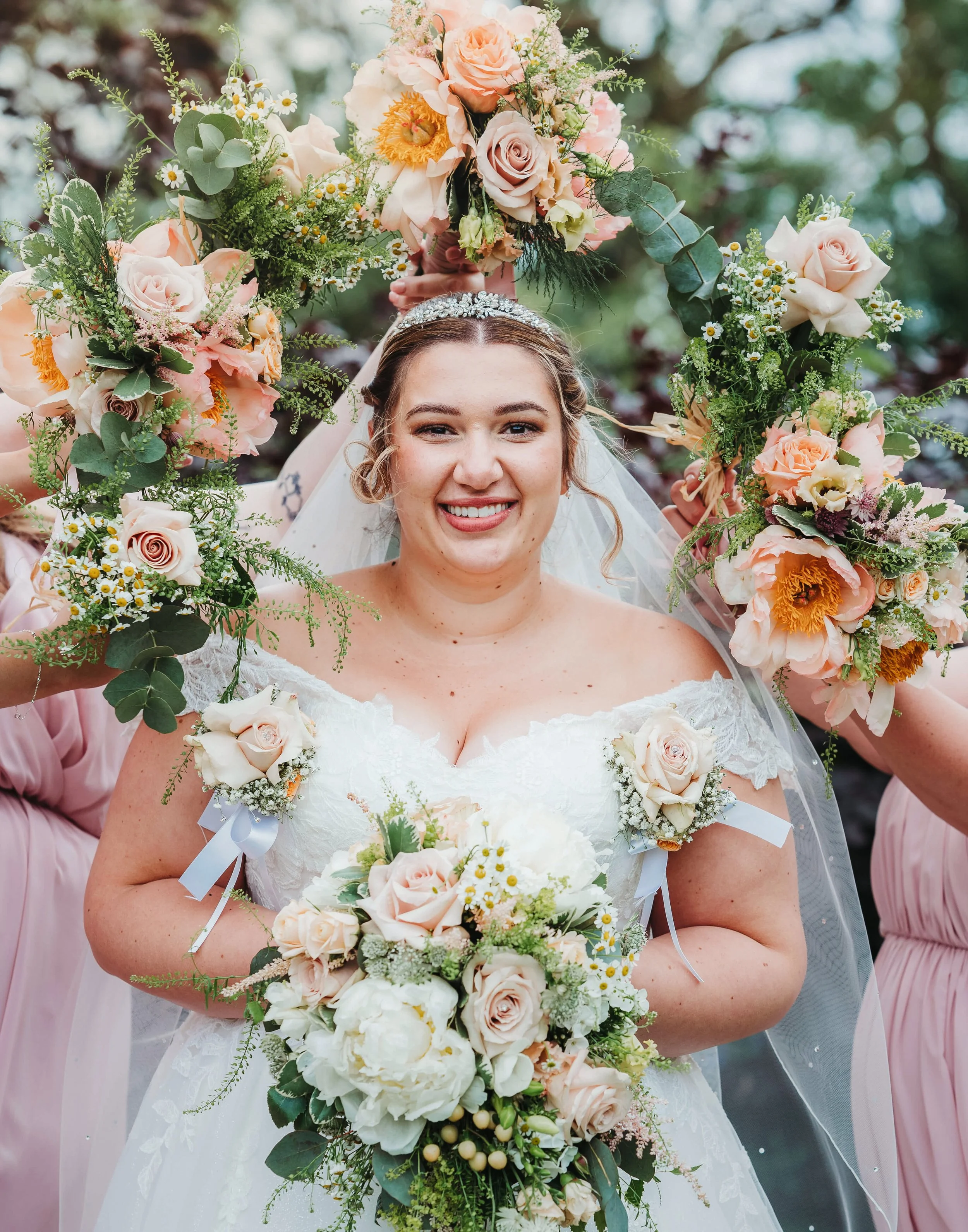 Bride smiling in a white wedding dress holding a bouquet of white and pale pink flowers, surrounded by friends holding a floral wreath overhead.