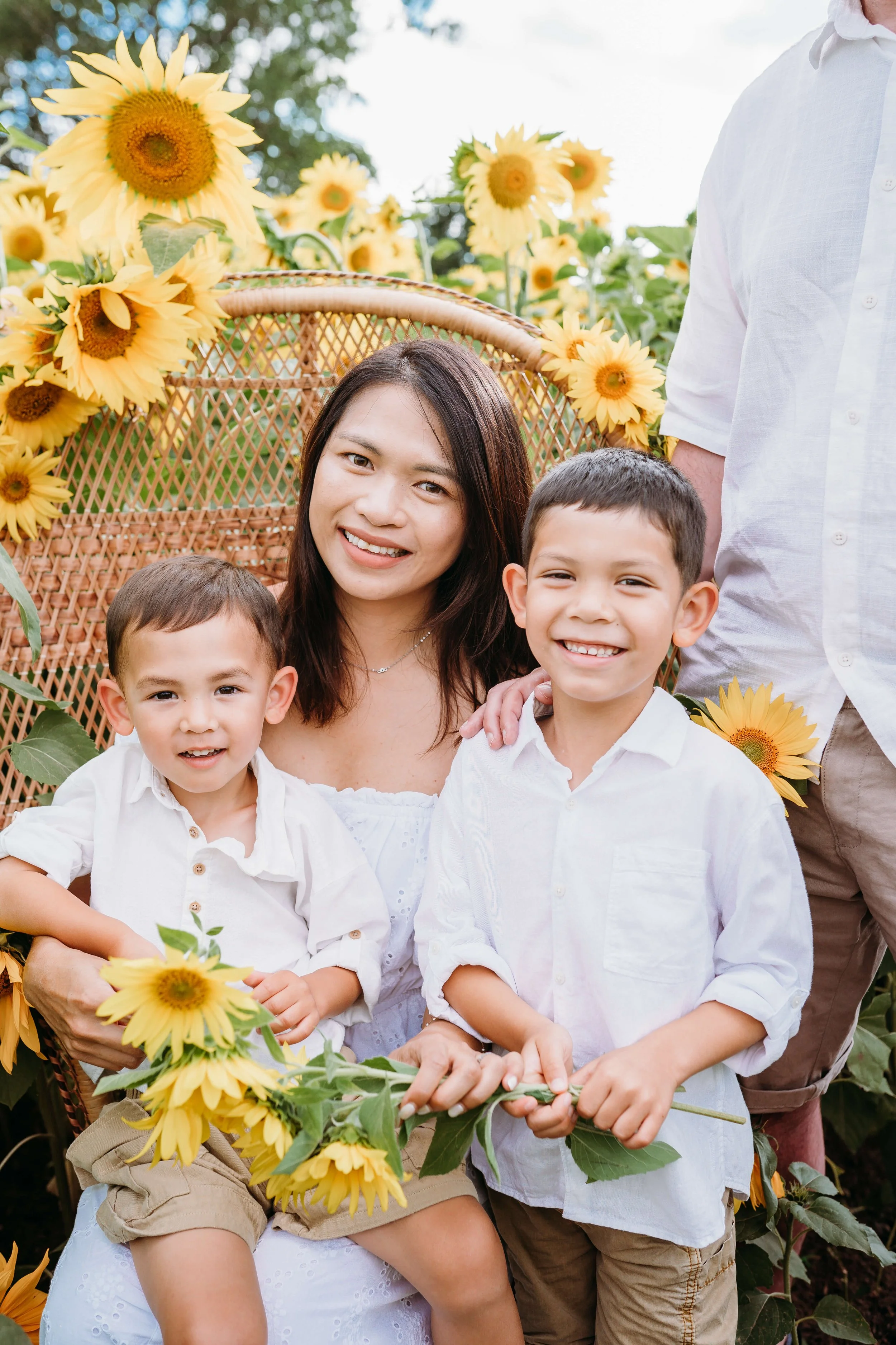 A woman and two young boys smiling in a sunflower field, with a wicker chair decorated with sunflowers behind them.