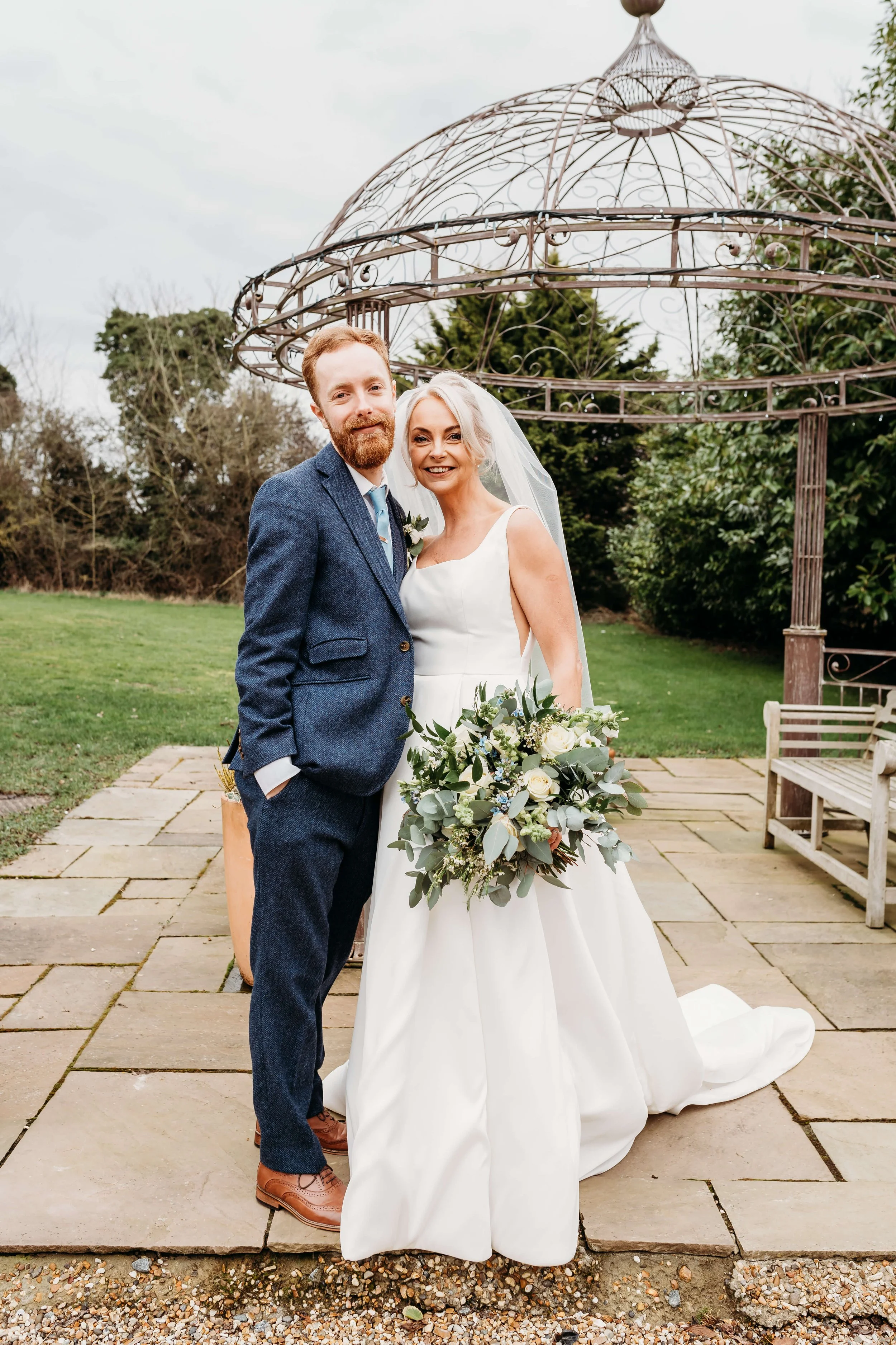 A bride and groom standing outdoors on a stone patio, smiling, with greenery and trees in the background. The bride holds a bouquet of white roses and greenery, and wears a white wedding dress with a veil. The groom wears a blue suit, white shirt, an