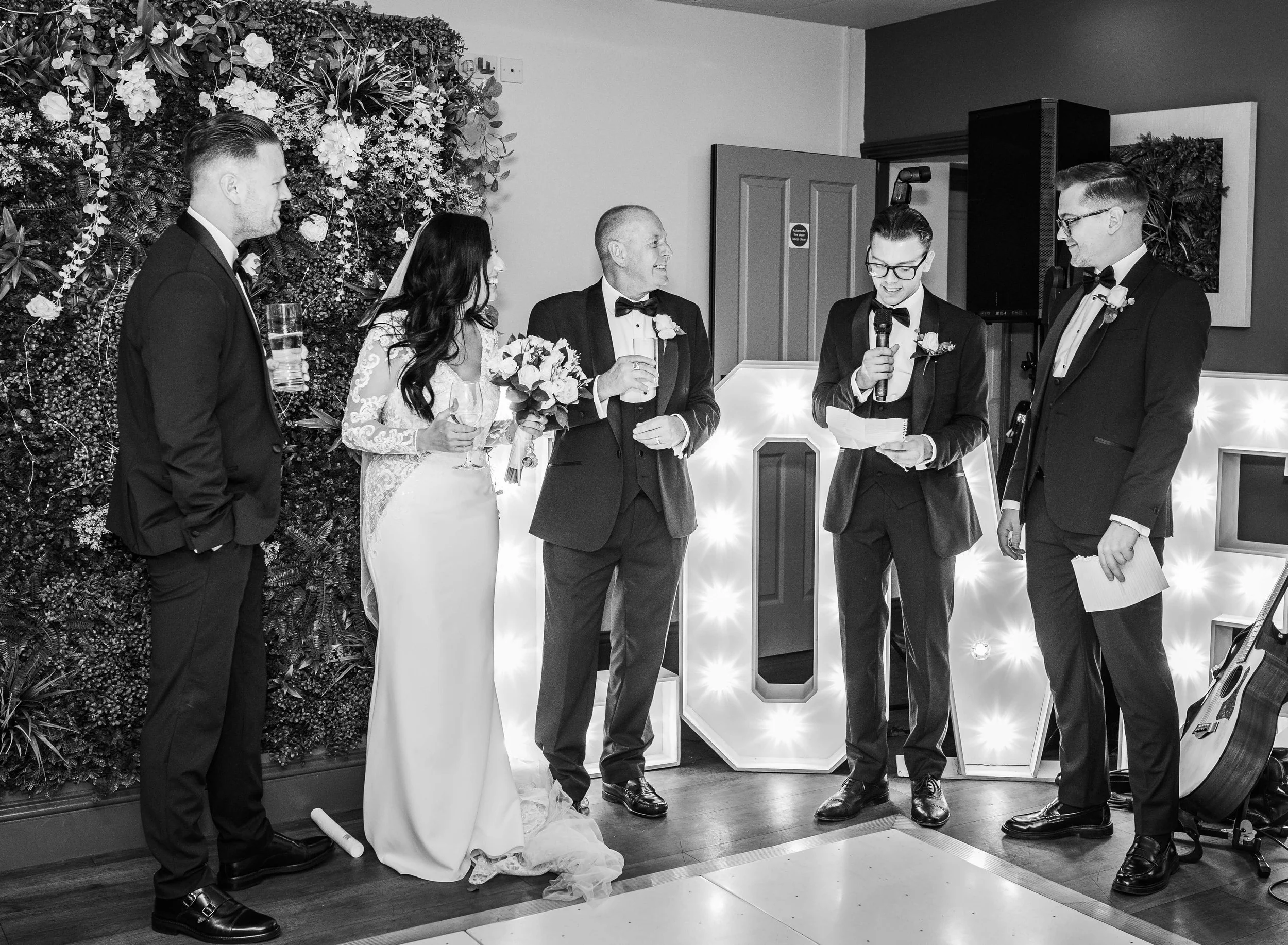 A wedding reception with six people, including the bride in a white gown holding a bouquet, and the groom in a black tuxedo. Others are in formal attire, with a person reading from a paper and holding a microphone, likely giving a speech or toast. Th