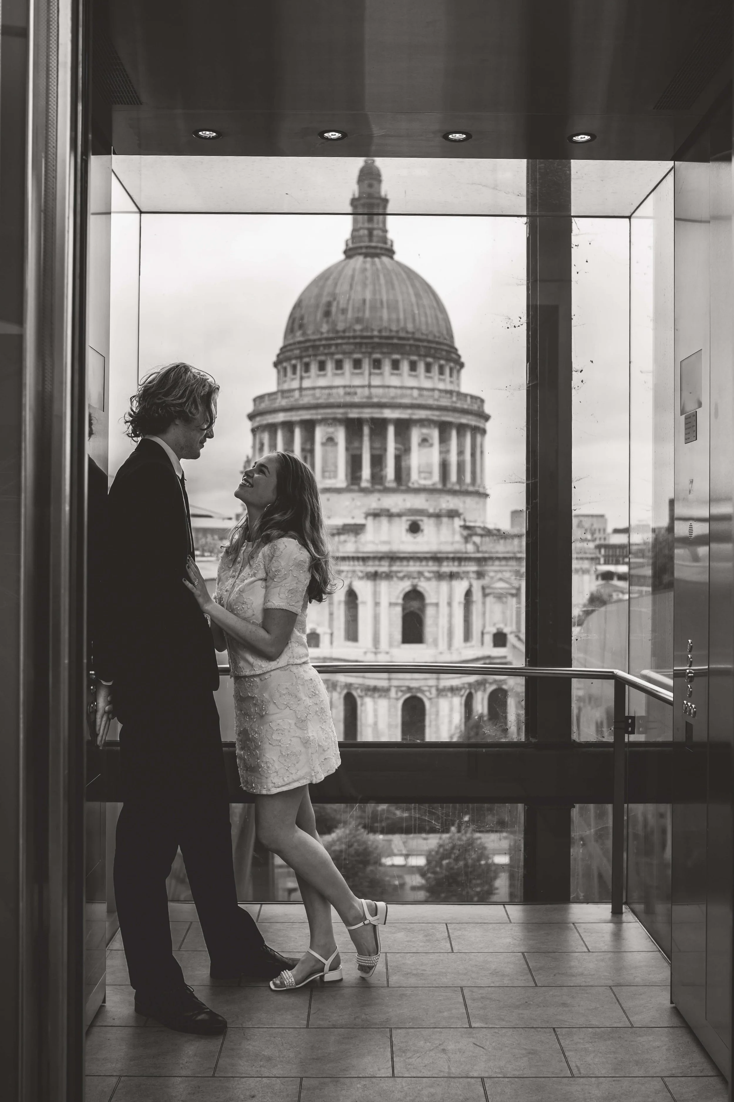 A black and white photo of a couple in an elevator, with the dome of St. Paul's Cathedral visible through the window behind them. The woman is wearing a dress and high heels, and the man is in a suit. They are looking at each other and smiling.