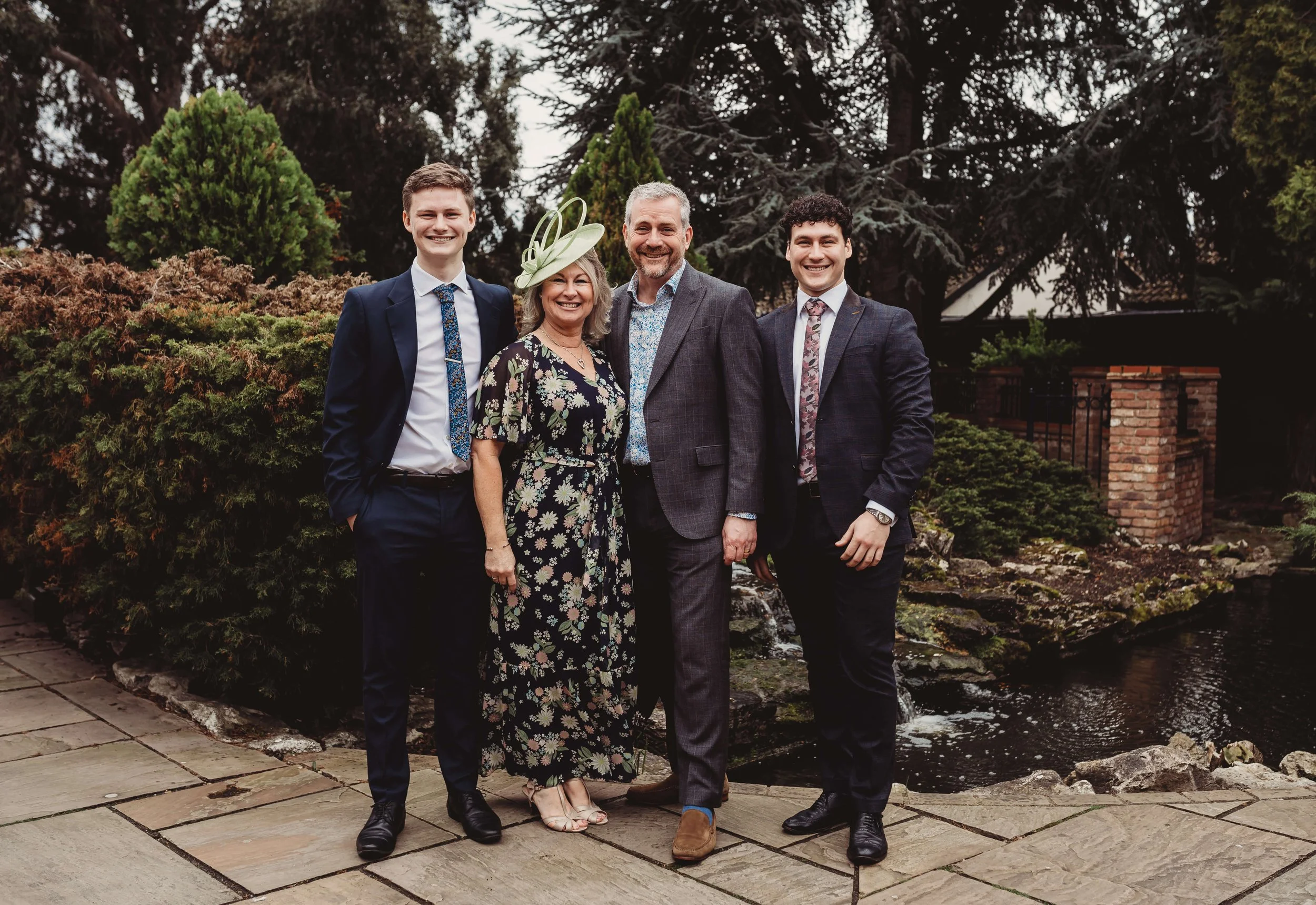 A family of five standing outdoors on a stone path, with greenery, bushes, and trees in the background. They are dressed in formal attire, smiling for the camera.