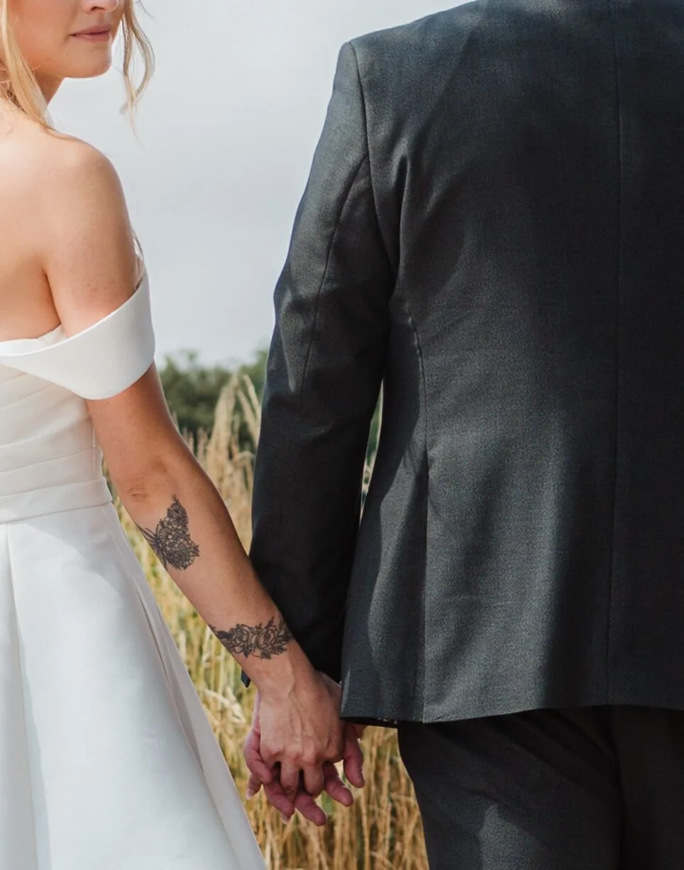 A bride and groom holding hands outdoors, with the bride wearing a white off-shoulder dress and the groom in a dark suit.