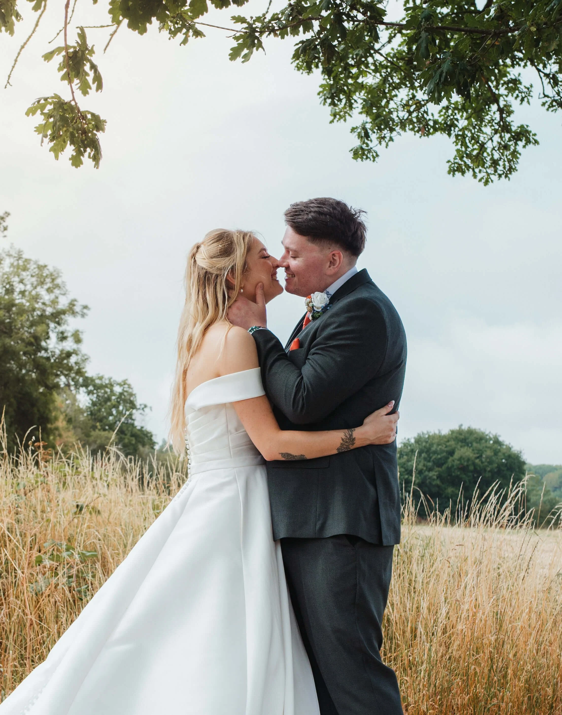 A bride and groom embrace and smile in a field, with trees and overcast sky in the background.