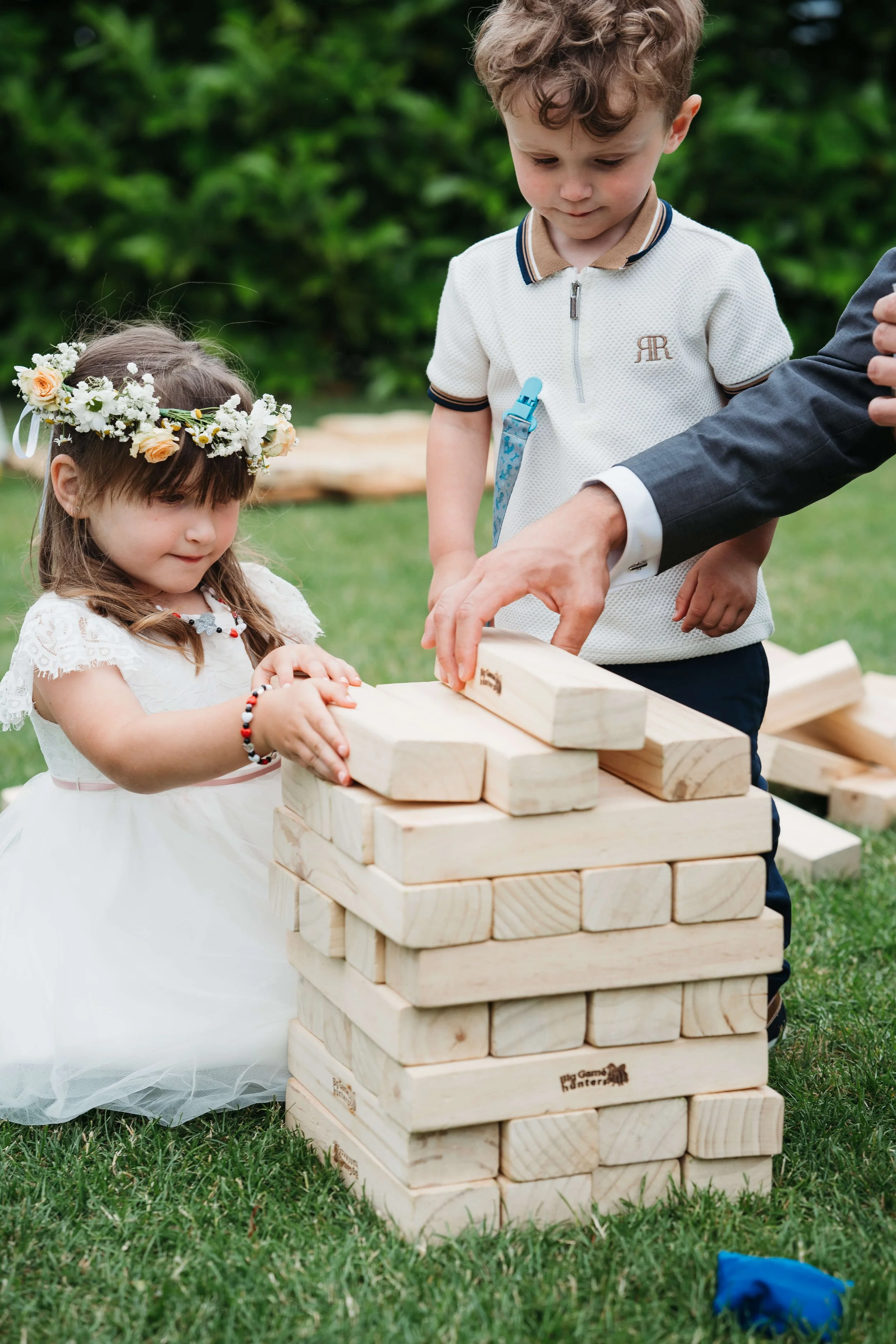 A young girl in a white dress and floral crown, and a young boy in a white polo shirt, play giant Jenga outside on grass with an adult.