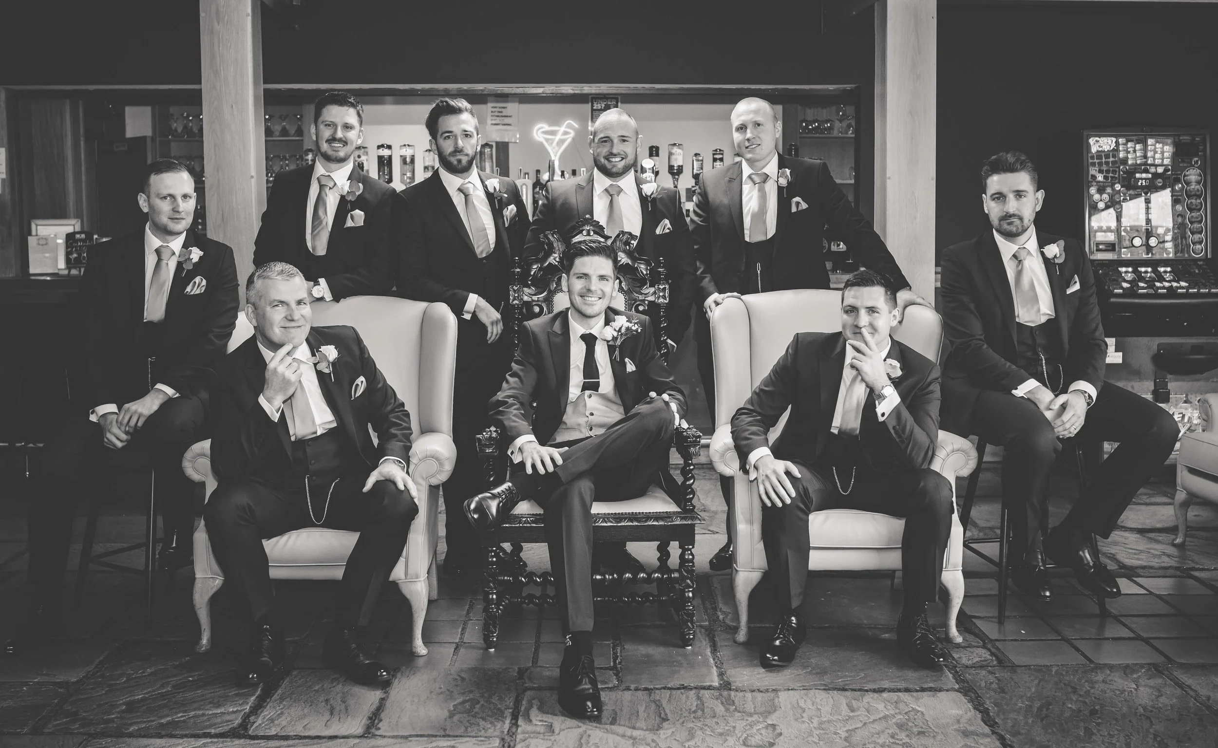 Black and white photo of ten men in suits, some sitting and some standing, in what appears to be a bar or lounge. The men are dressed formally, with some seated on chairs and others standing behind. The setting includes a bar counter with bottles, an