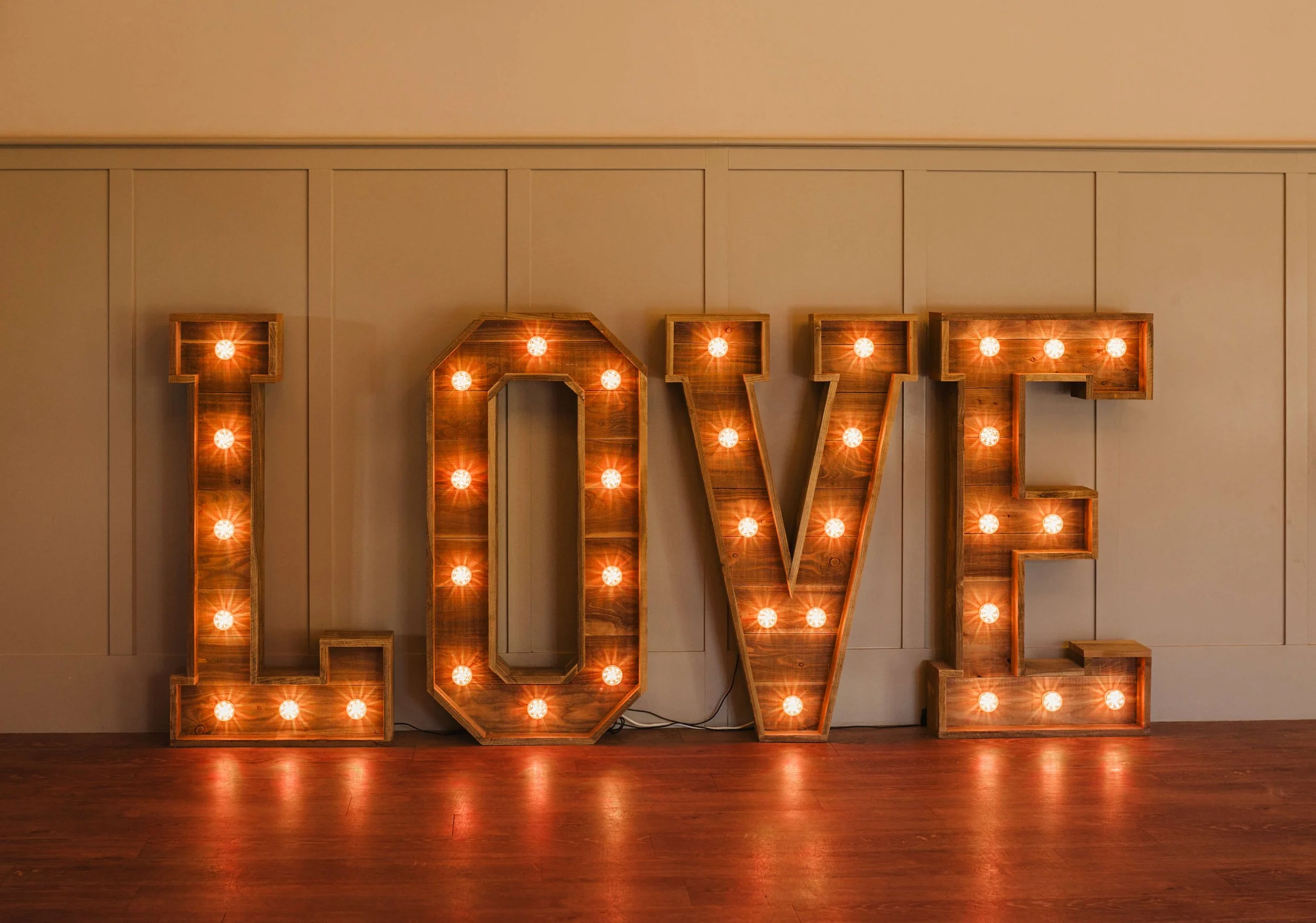 Large illuminated letters spelling out 'LOVE' with warm light bulbs inside, placed against a beige wall on a wooden floor.