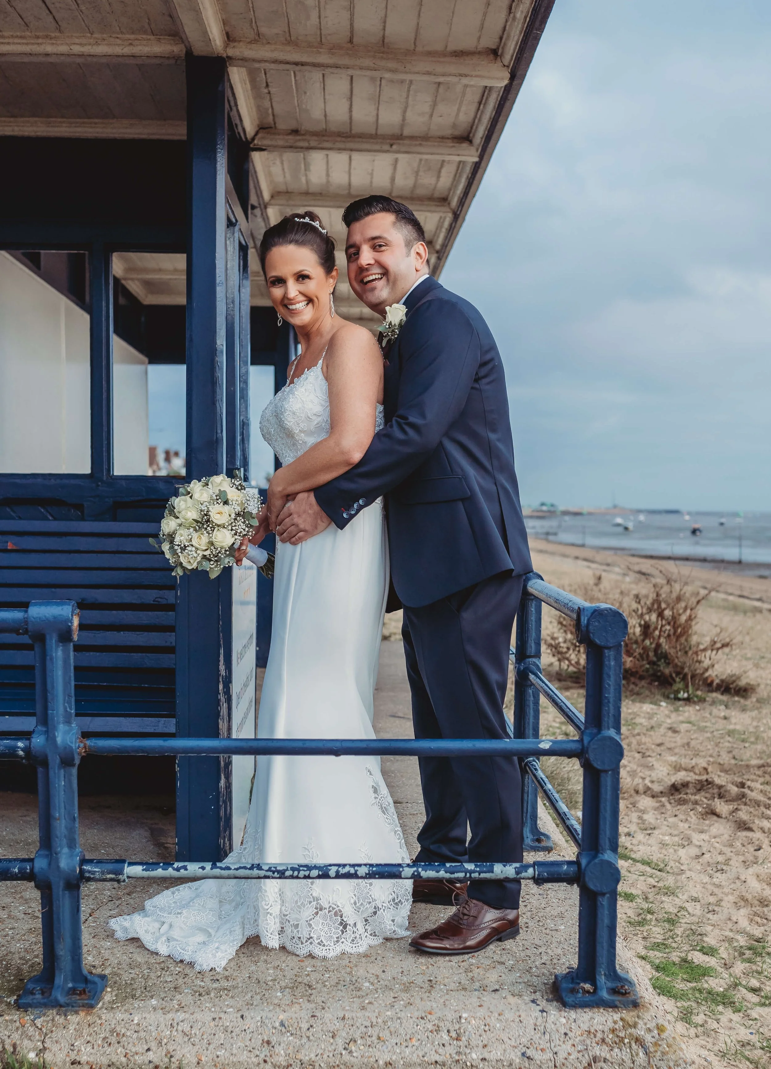 A newlywed couple smiling and holding a bouquet, standing on a beachside structure with the ocean in the background.