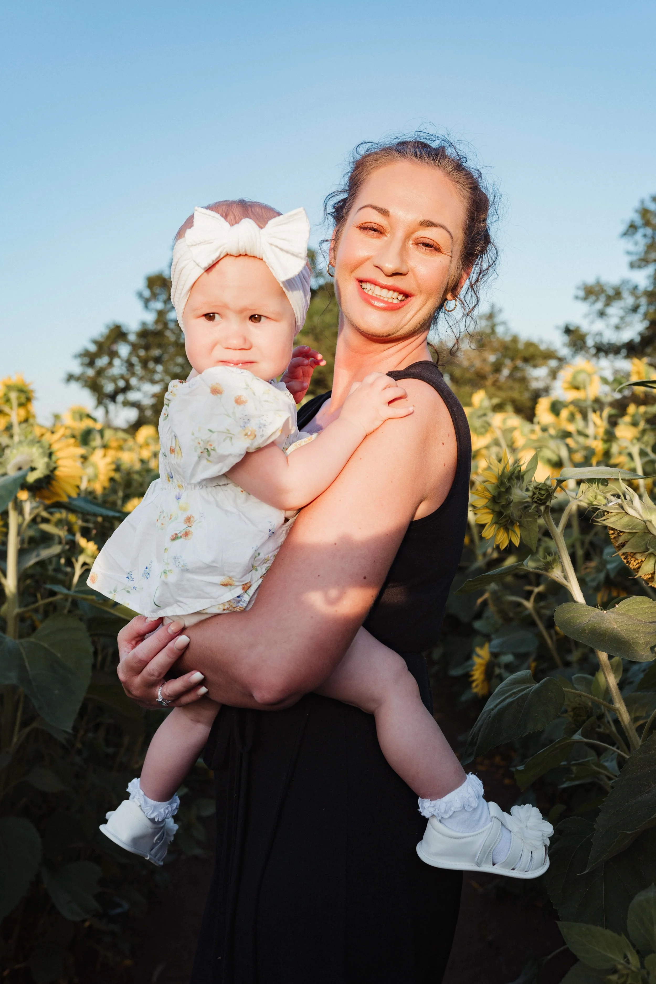 A smiling woman holding a young girl in a sunflower field during sunset.